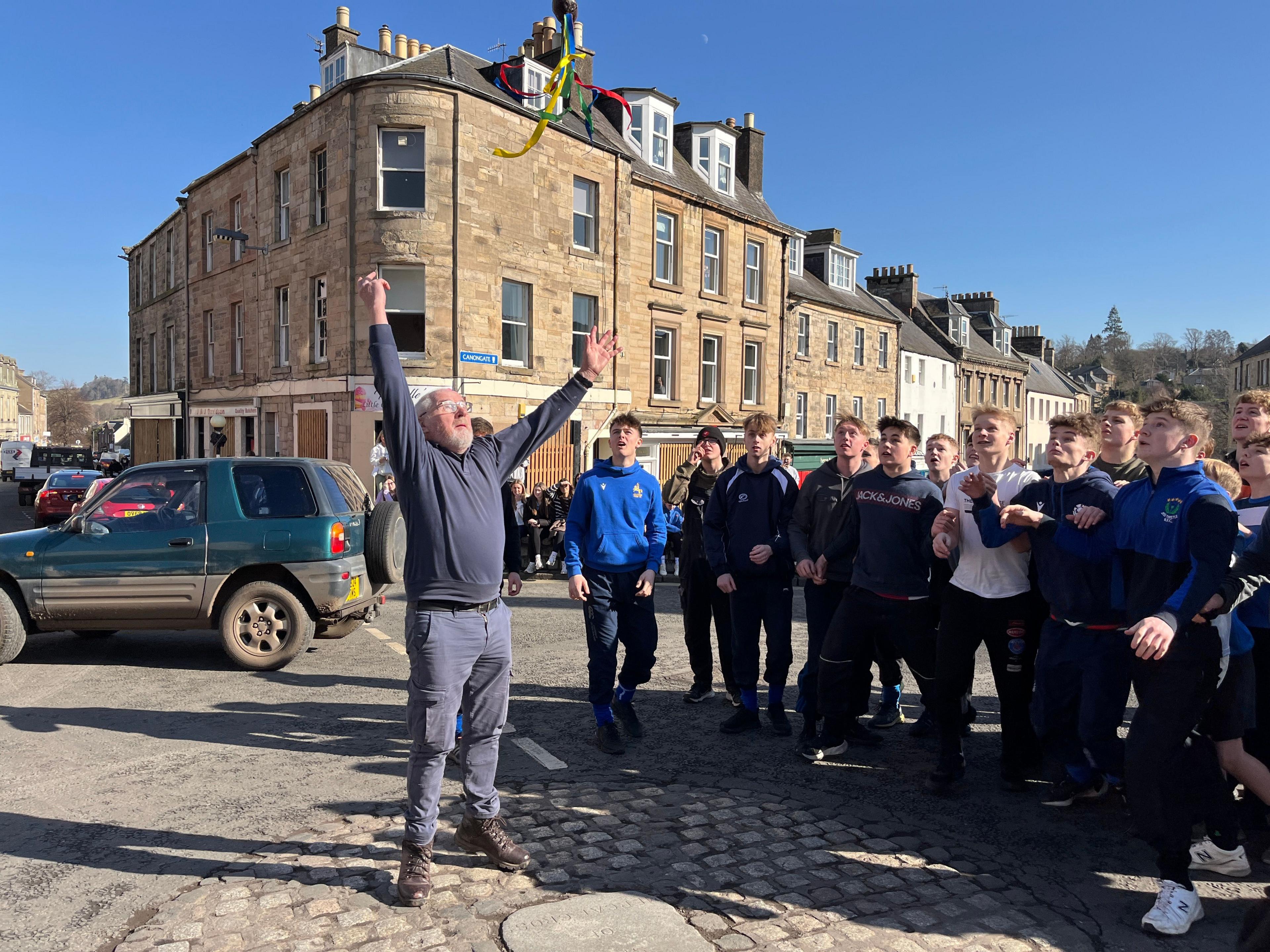 A man throws a small leather ball into the air to get the Jedburgh ba' games under way. A group of young men have gathered next to him on the road. 