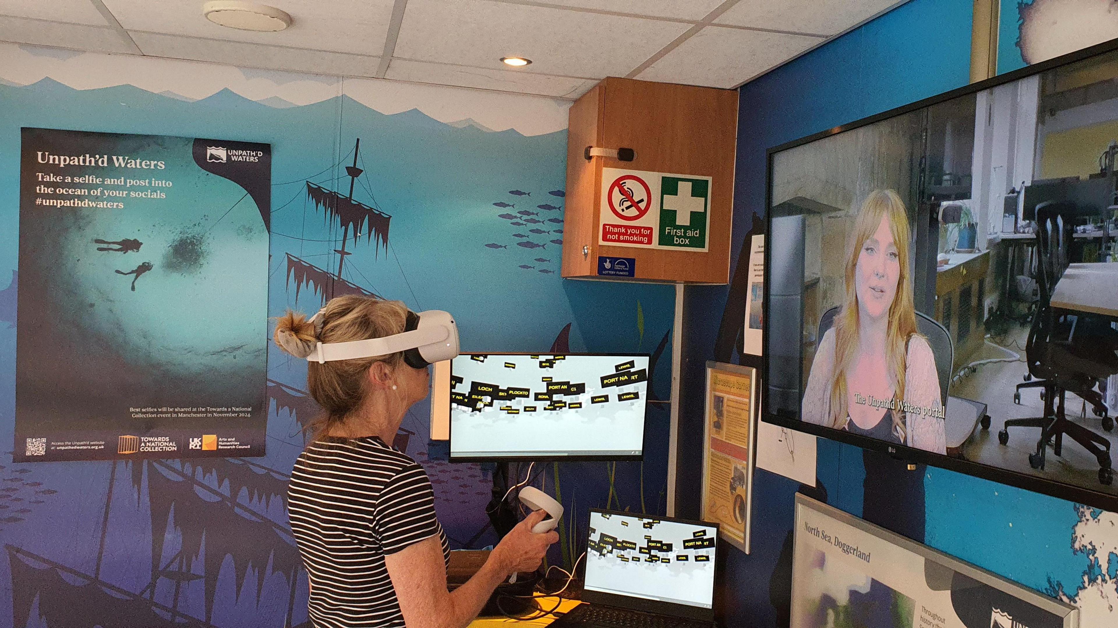 A woman wearing virtual reality goggles with a controller in room with a blue background, and screens on the wall.