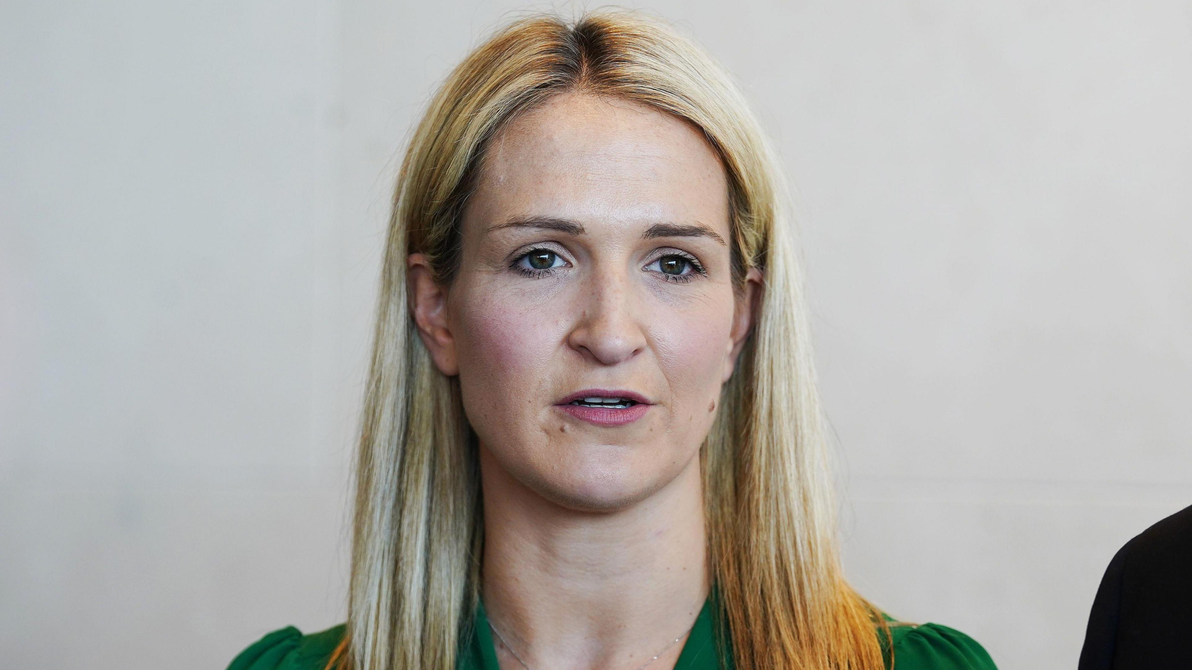 Helen McEntee - a woman with long, blonde hair wears a green dress. She is standing in front of a white wall with her mouth slightly open like she is talking to someone behind the camera.