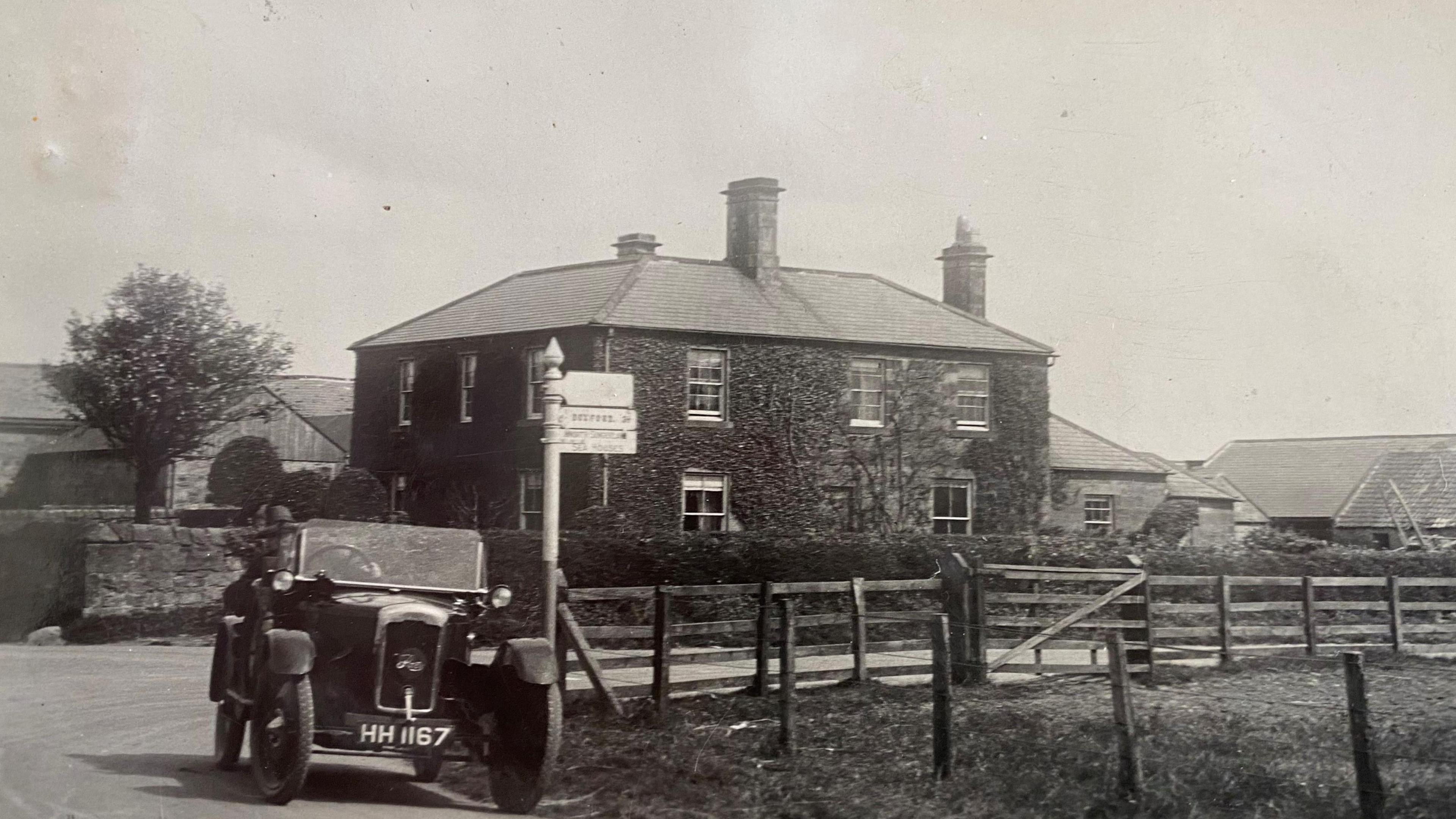 A black and white image of Charlton Mires taken in the 1920s with a car of the era being driven outside. The house is a large Georgian one with three chimneys and there are farm buildings behind and a wooden fence in the foreground. It sits on a junction with an old road sign.
