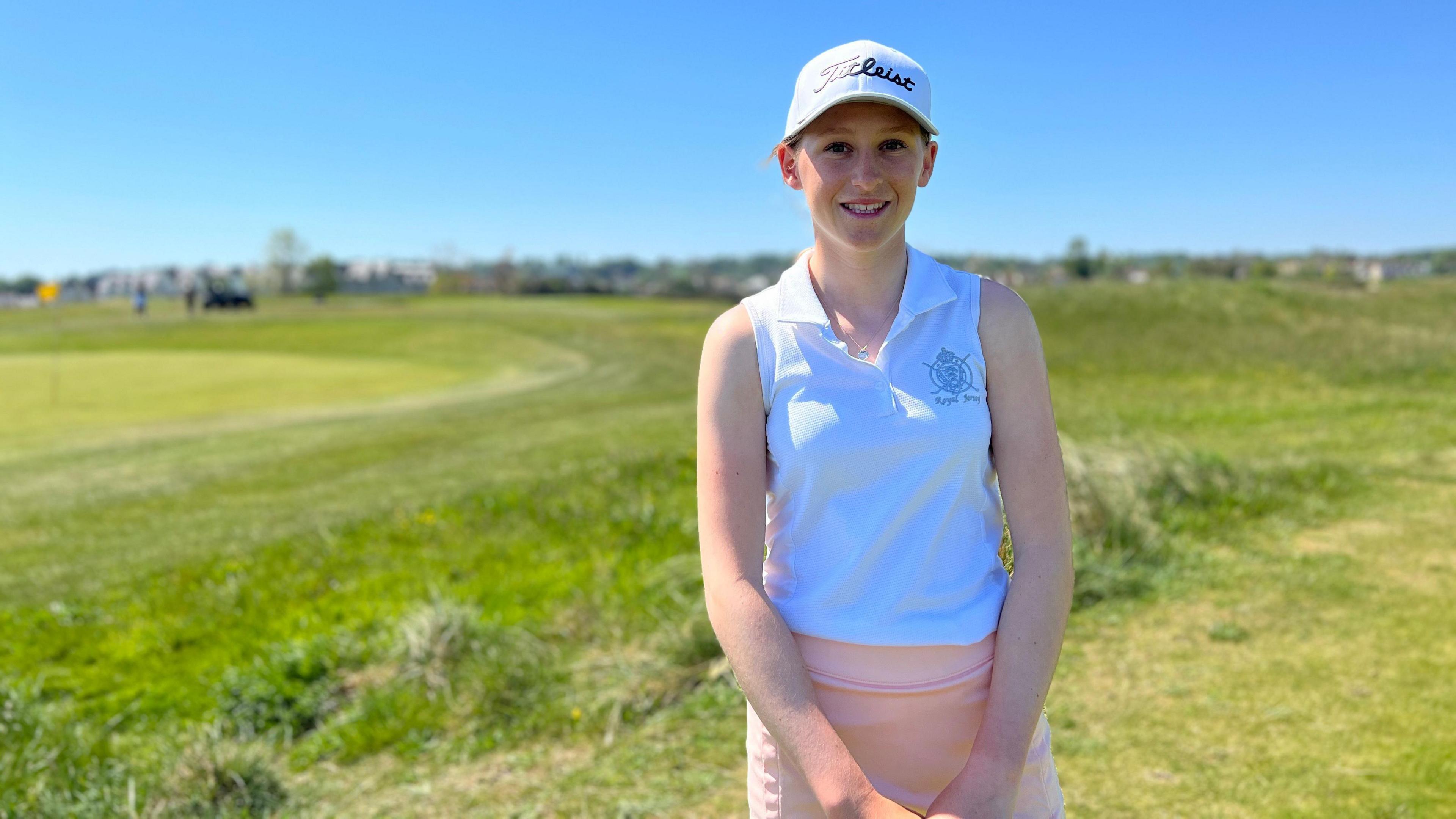 Anabelle Lucas-Villar smiles at the camera as she wears a pink skirt and white sleeveless top and a white cap. Behind her is a golf course.
