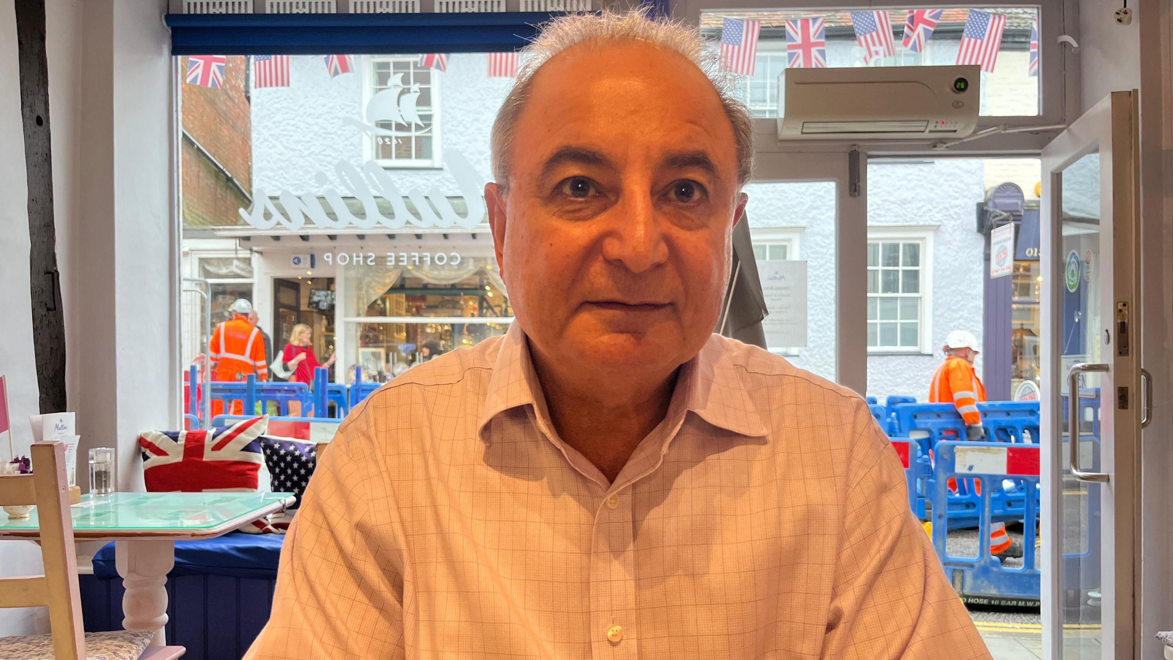 Basil Doha sits in Mullins Coffee Shop in a shirt, looking straight at the camera. Behind him is a large window and the street and roadworks can be seen outside.