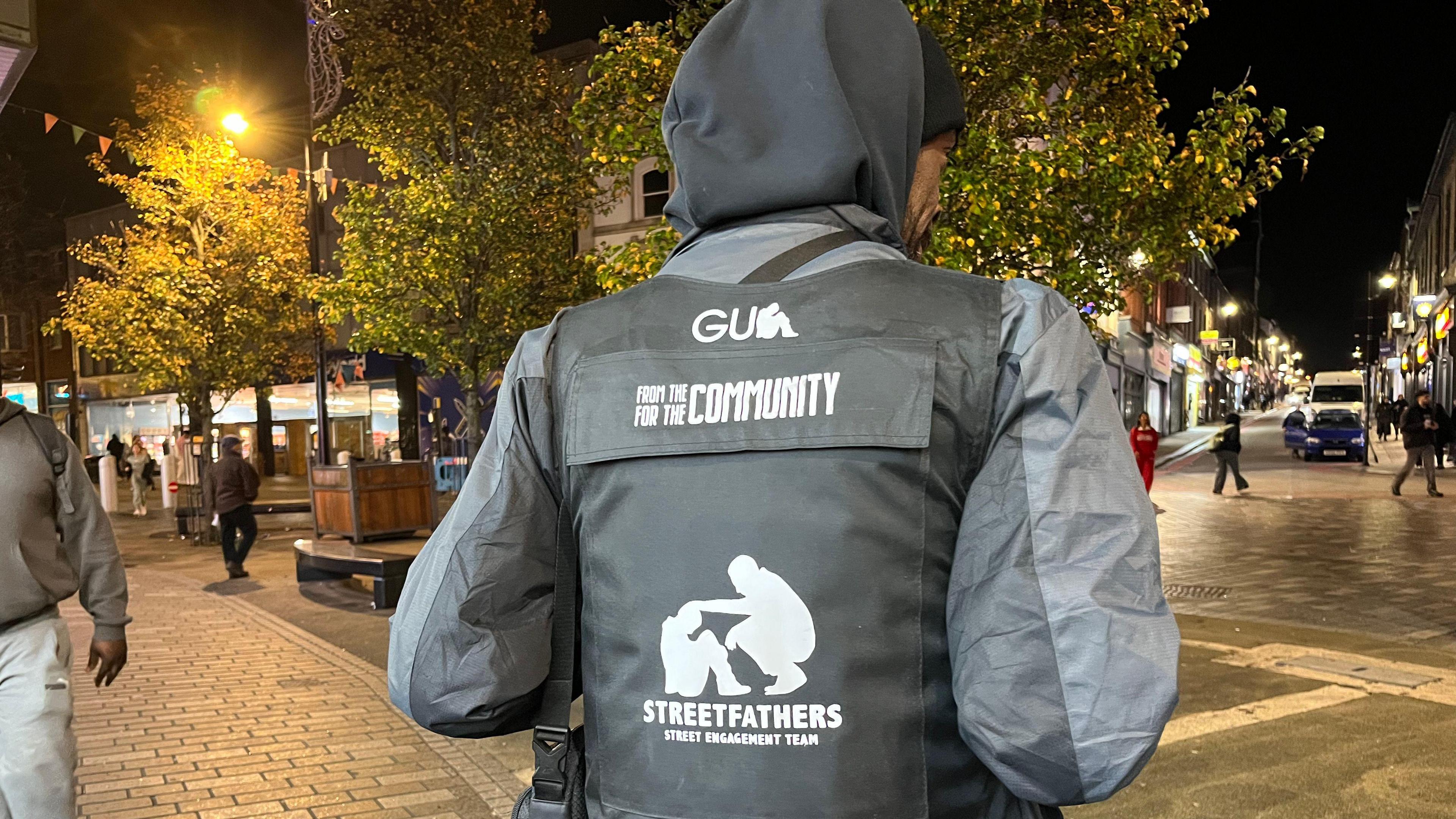 A person is standing on a street at night, wearing a dark hooded jacket and a black stab-proof vest. The vest has white text and graphics on the back. At the top, it says “FROM THE COMMUNITY FOR THE COMMUNITY.” In the centre, there is a silhouette graphic of an adult crouching and placing a hand on the shoulder of a child. Underneath, the words read: “STREETFATHERS – STREET ENGAGEMENT TEAM.”