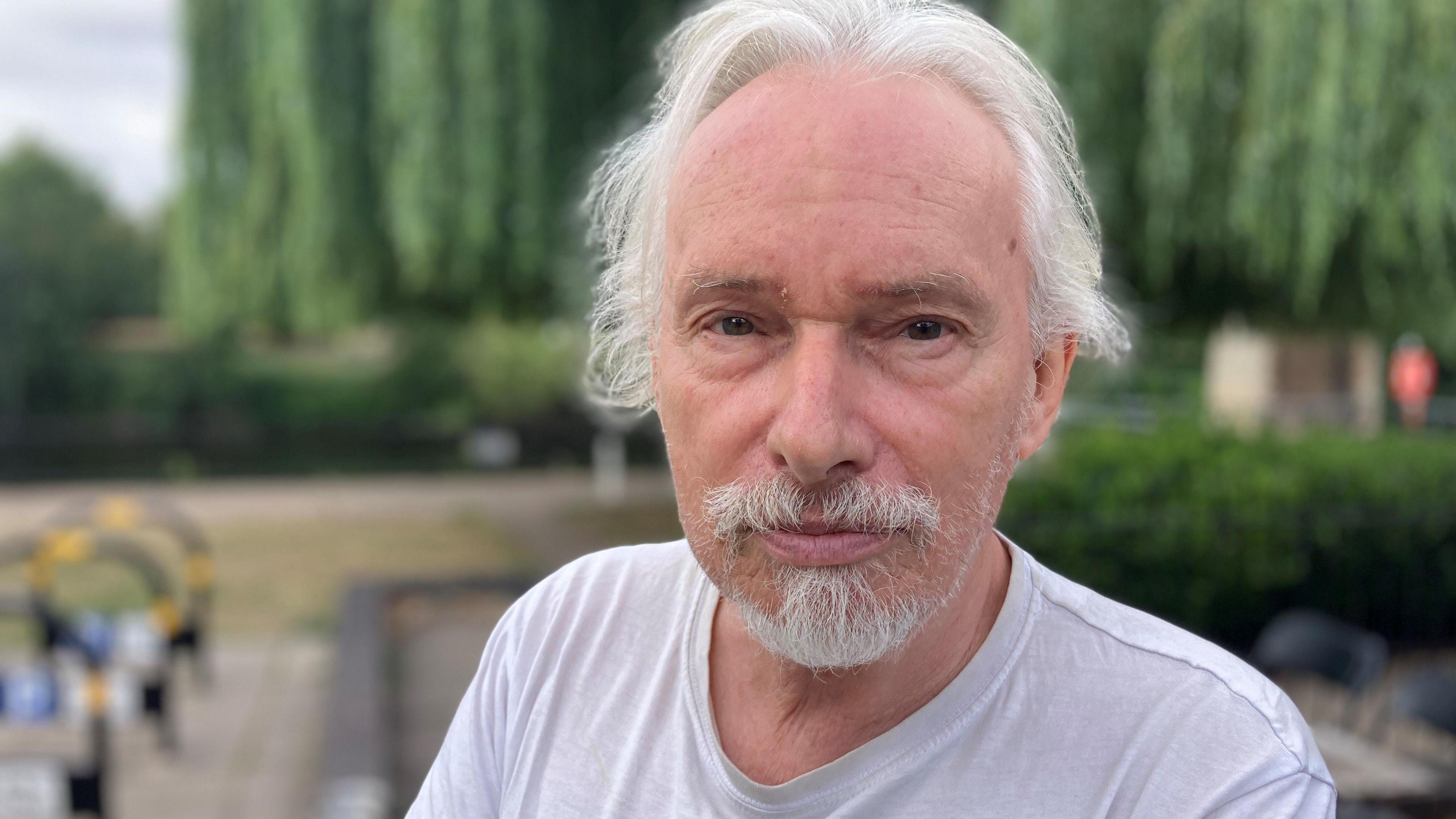 With white hair, white goatee and white T-shirt, Stephen Pain stands next to the river Severn in Worcester's Riverside park. Cycle racks and a table and chairs are visible in the background