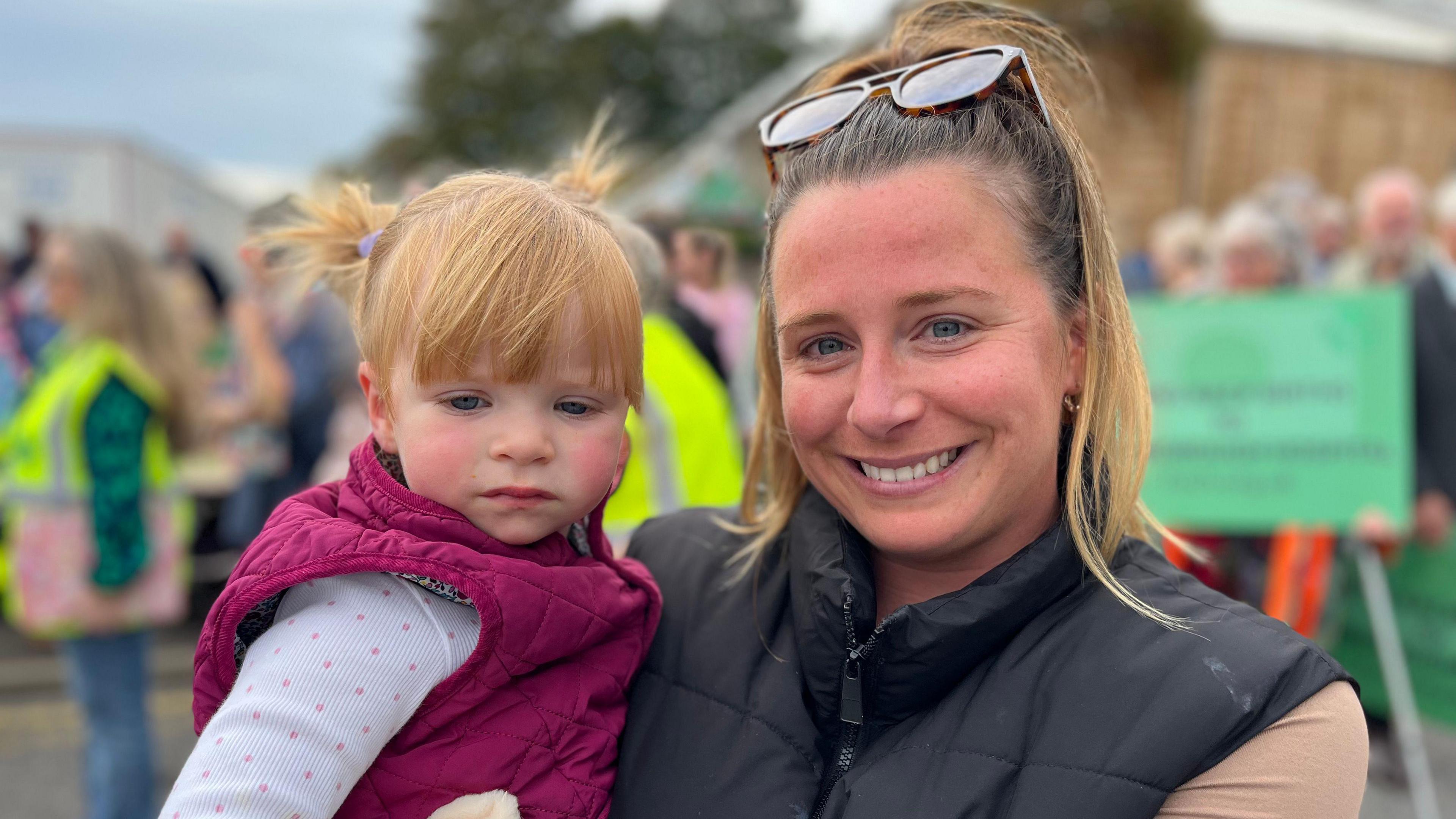 Lucy humphrey with her daughter in her arms, a crowd holding placards are behind them. They both have strawberry blonde hair and lucy has sunglasses on her head