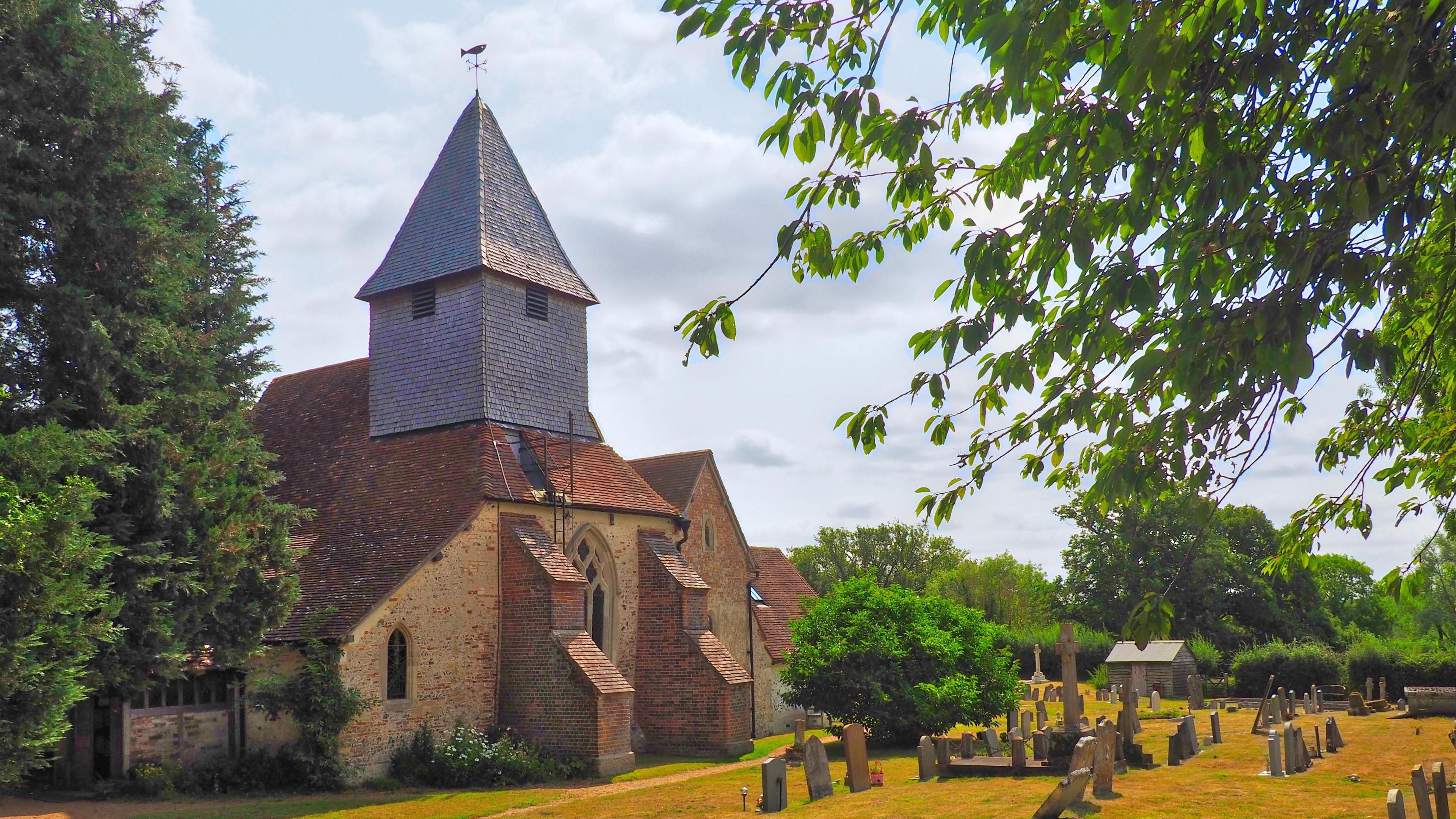 A stone and brick church with a grey tower stands between two green trees at Silchester. There are several grey gravestones in front of the church in short dry yellowing grass. There are several green trees dotted throughout the picture.