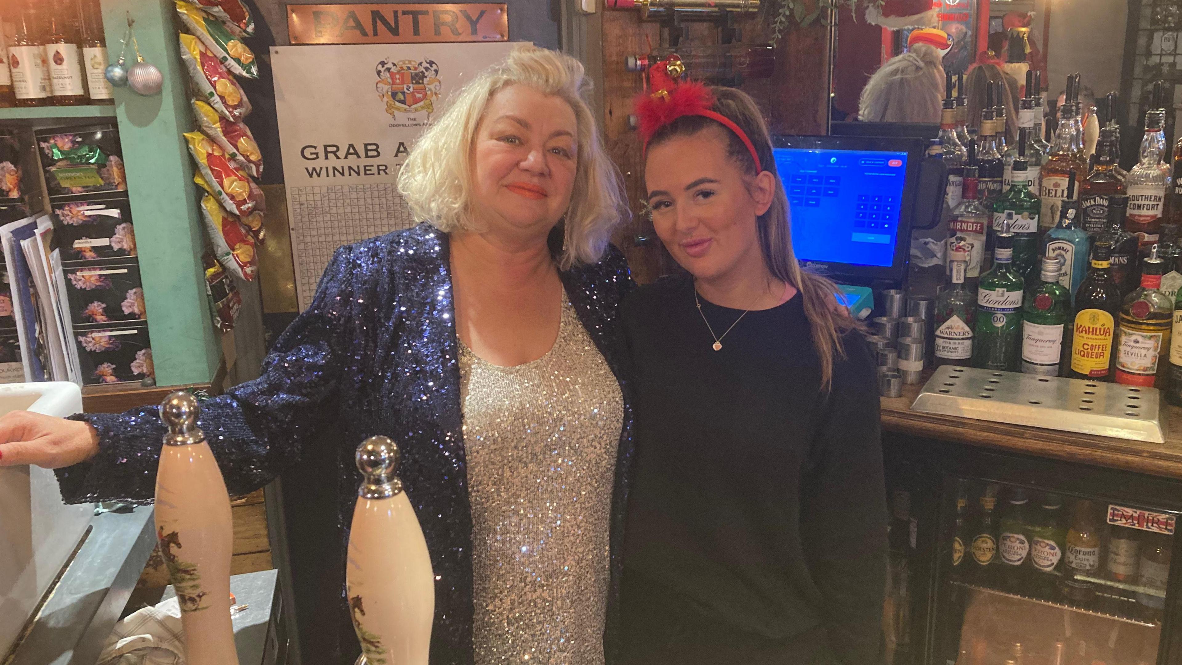 Niki Hackett is dressed in a blue glittery cardigan and a silver glittery top. She is stood behind the bar with a younger member of staff dressed in black and wearing a Christmas themed headband.