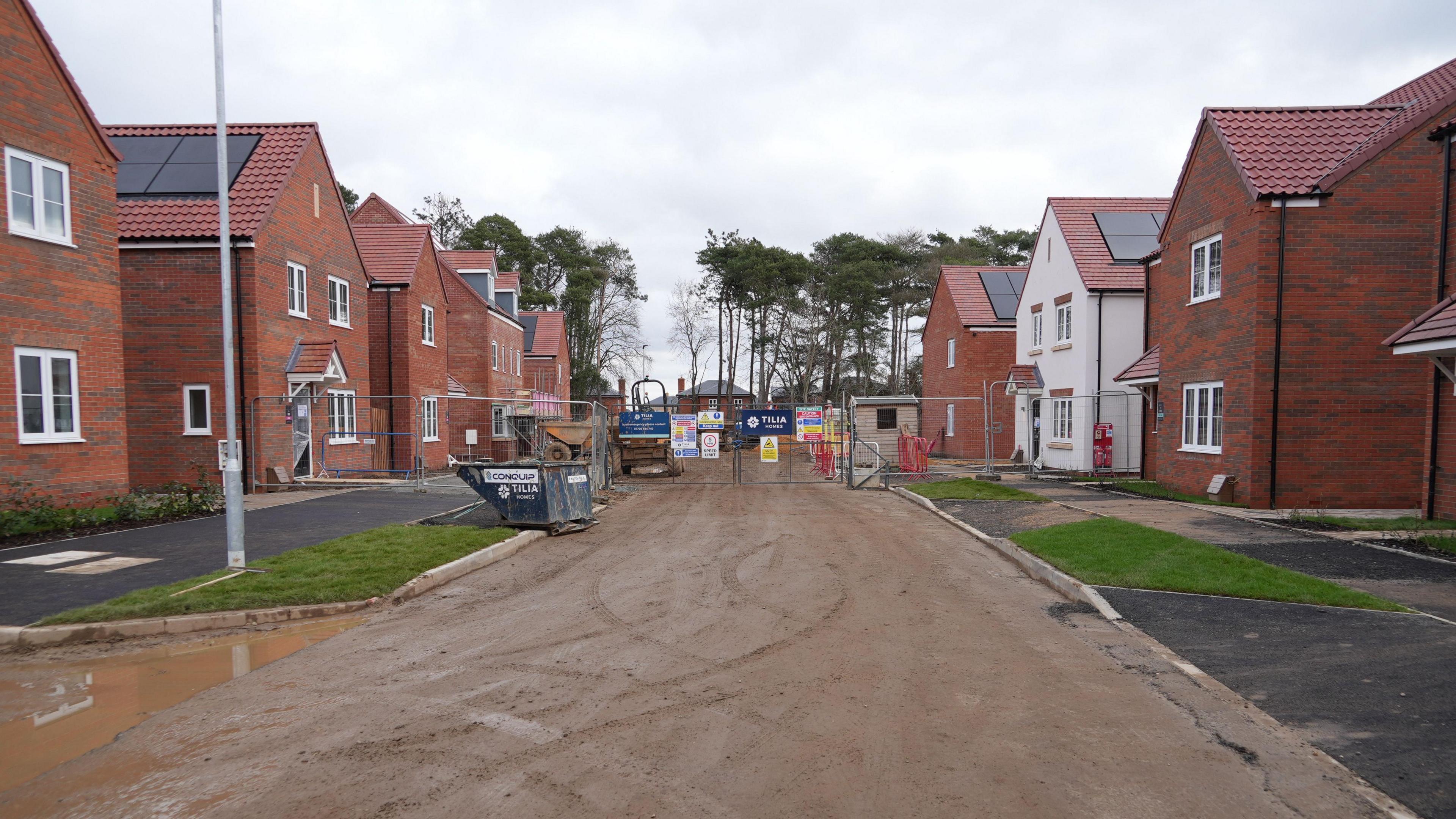 Metal fencing, with construction notices block off a muddy road. There are new build houses in front and behind the fencing.