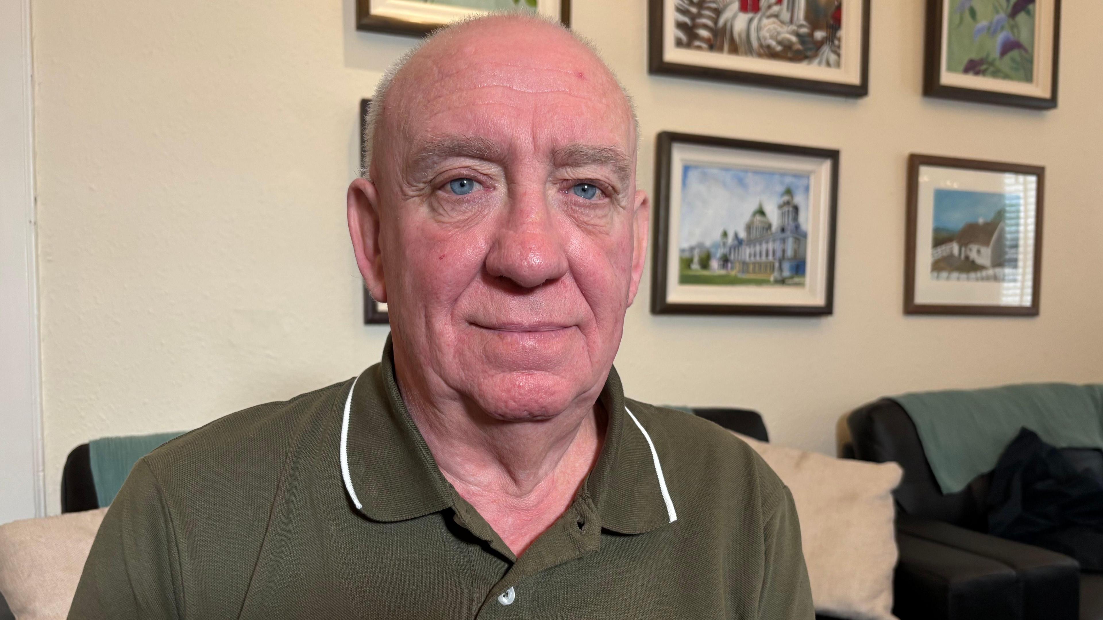 Alex Bunting is sitting in a room which has framed pictures on the wall. He wears a green T-shirt.