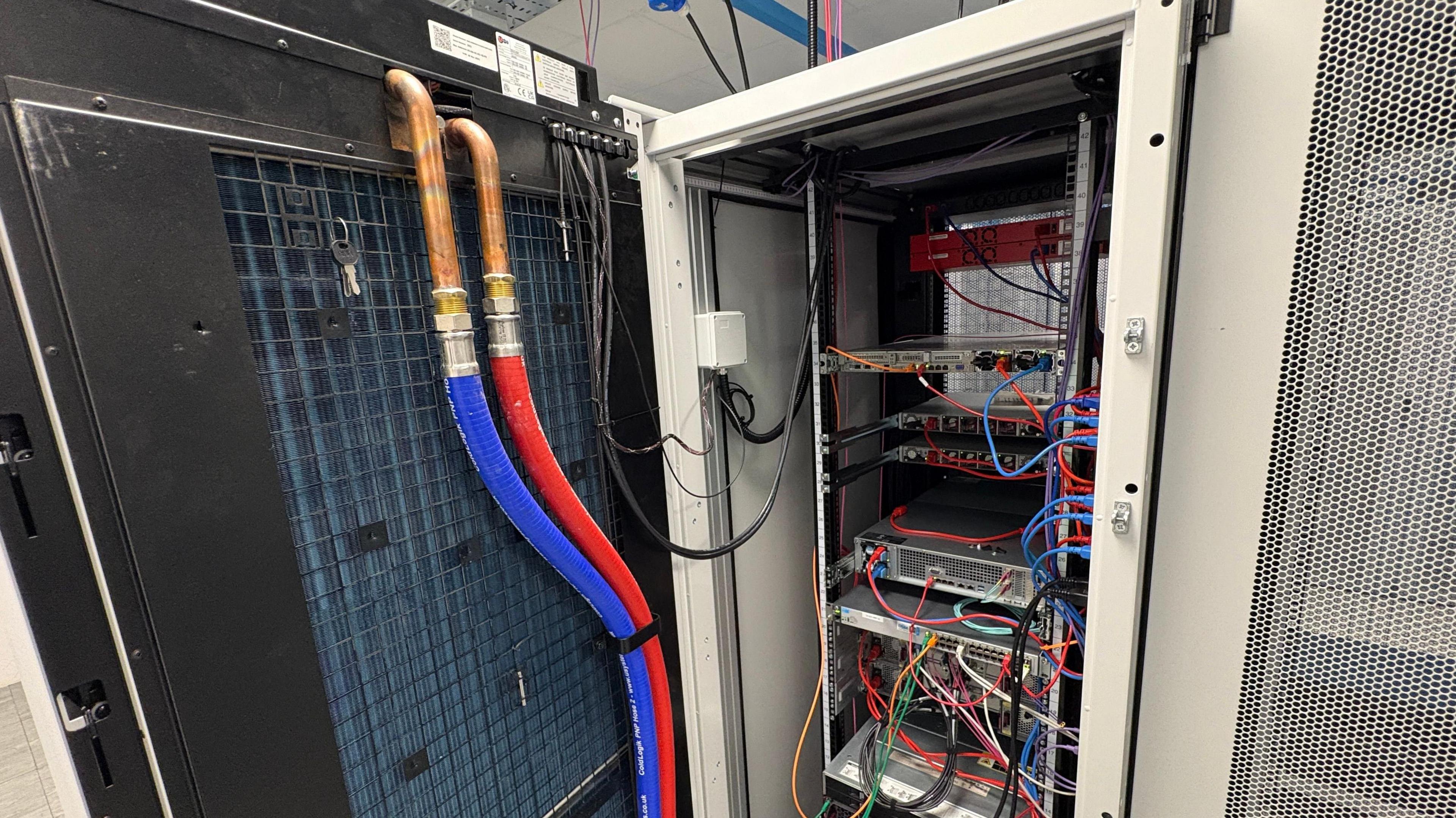 A view inside a server rack at DSM's lake-cooled data centre. It is a high-tech cupboard, the door of which has been opened and stands ajar on the left of the image. Two pipes – one red and one blue – are running roughly vertically up the inside of the open door. They each connect to two copper pipes attached to the door. Under the pipes is a black metal grille. Inside the cupboard are shelves containing computing equipment and wires.
