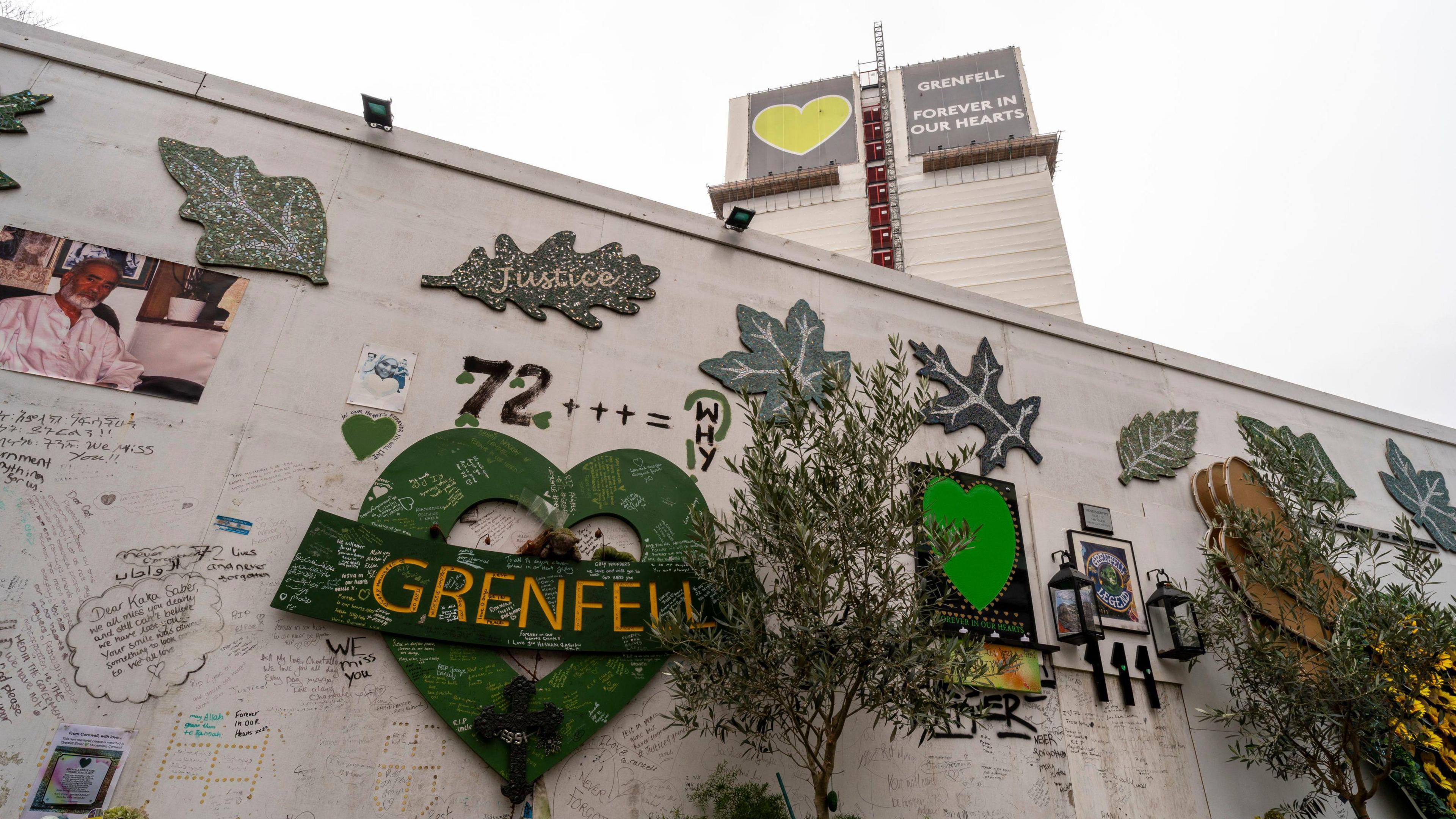 A file image showing Grenfell Tower covered in tarpaulin and a grey and green sign with a heart saying 'Grenfell: Forever in our hearts' in the background while white hoarding sits in the foreground. A green heart with the word 'Grenfell', green leaves, picture and handwritten tributes cover the hoarding