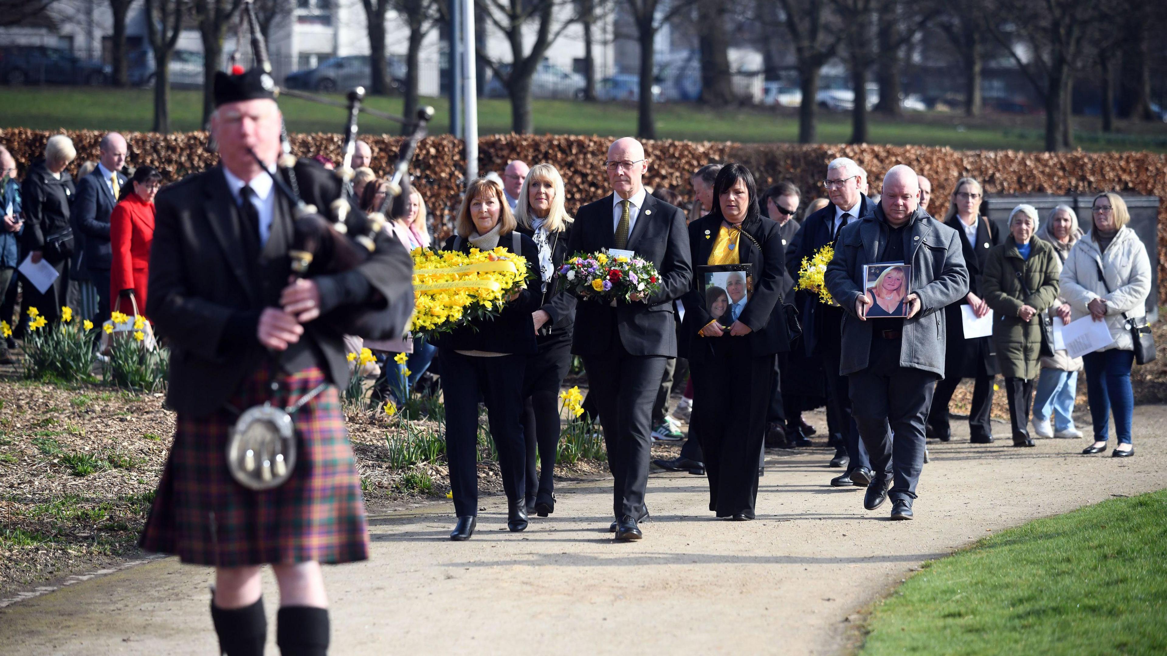 A man in a  kilt playing the bagpipes, with John swinney and a small crowd of people walking behind.