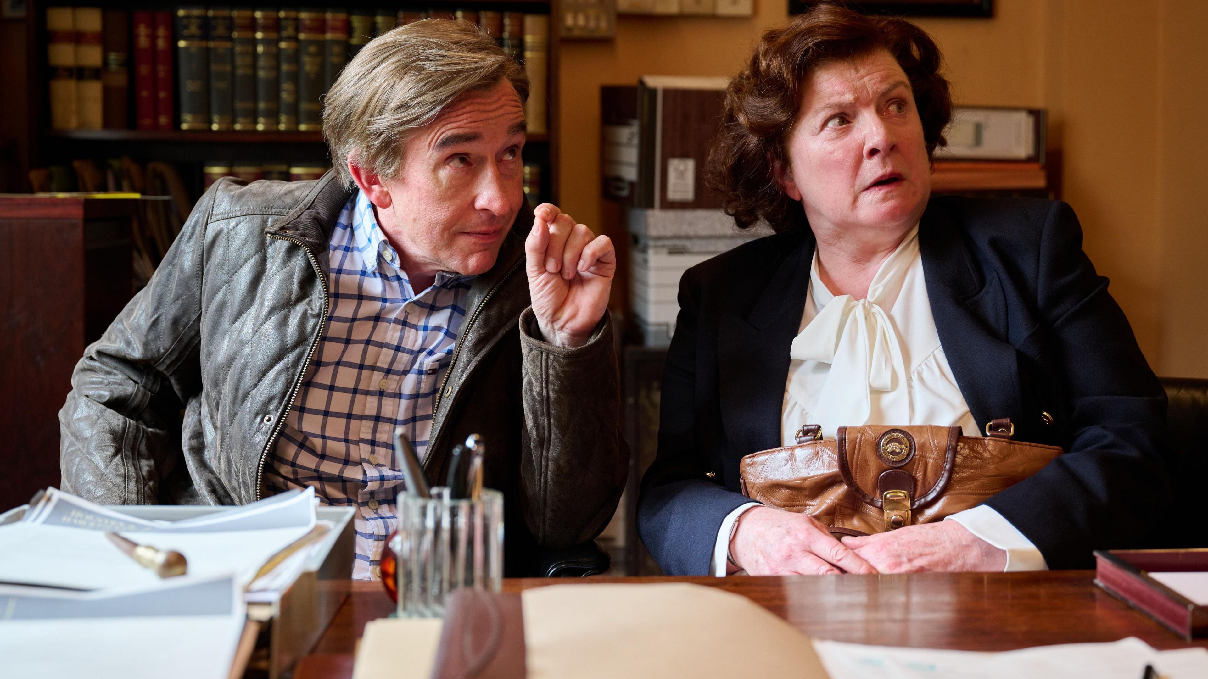 Steve Coogan as Alan Partridge sits at a desk looking over it to the side. He has his hand up and is wearing a checked shirt and padded leather jacket. He is looking hopeful. Lynn is sat next to him, clutching a handbag on her lap. She is wearing a navy jacket and a cream with a pussy bow. She is looking surprised.  Photo strictly embargoed by production company BBC until Fri 3 October.