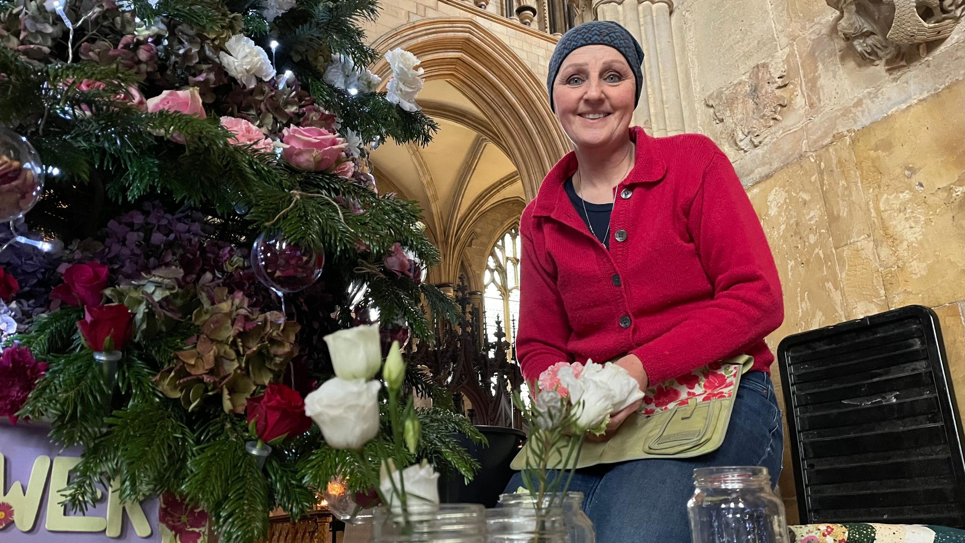 A fir Christmas tree on the left of the picture is decorated with white carnations, pink and red roses and burgandy hydranges. Clear baubles hang on the tree, containing dried dark red rose petals. A woman wearing a dark blue, patterned headscarf, red cardigan and blue jeans is kneeling next to the tree. She is looking at the camera and smiling.