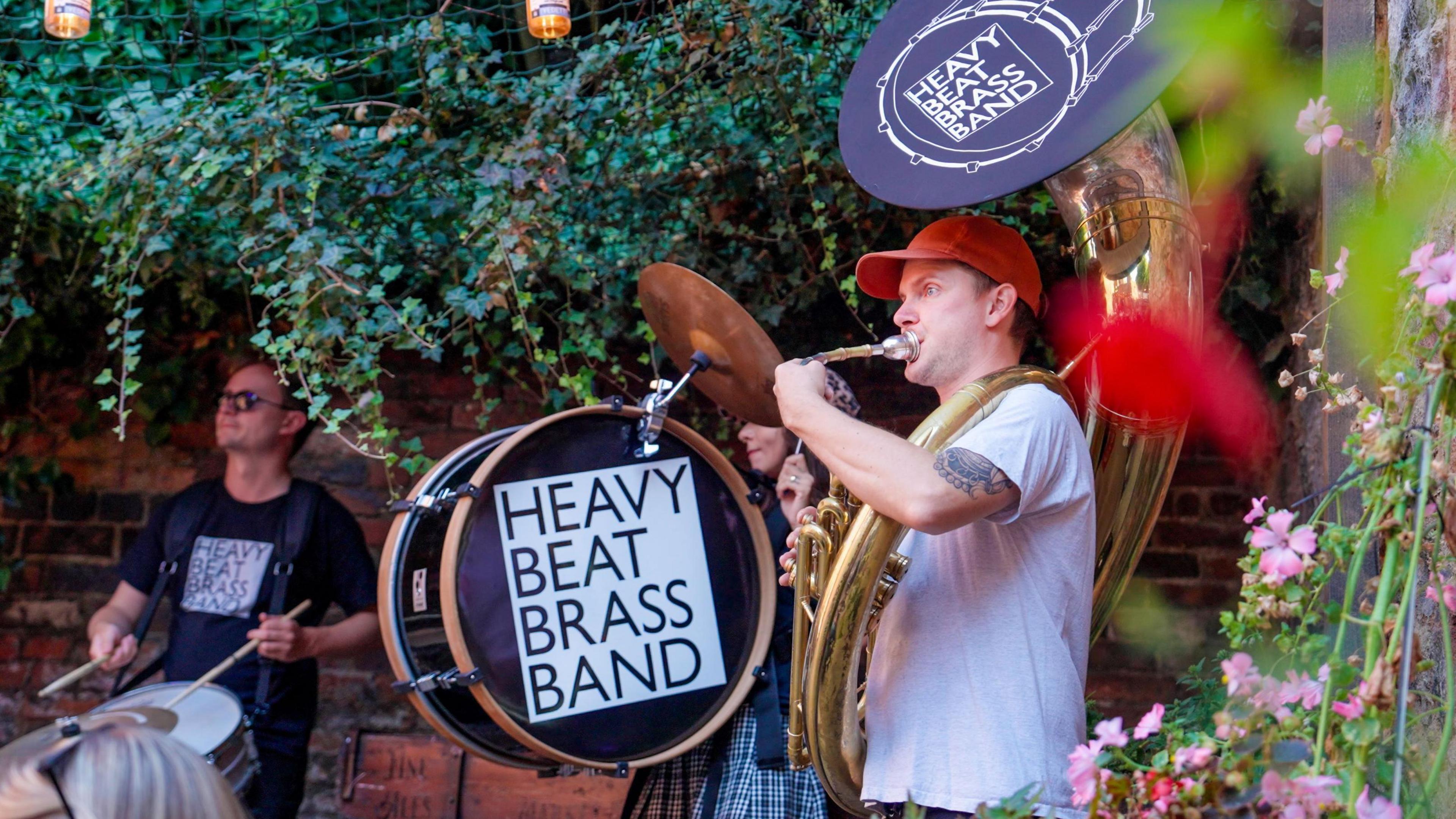 Members of a band, one of whom is carrying a bass drum with a sign saying 'Heavy Beat Brass Band". A man in a red cap is playing a brass instrument and a man in the background is playing a drum. 