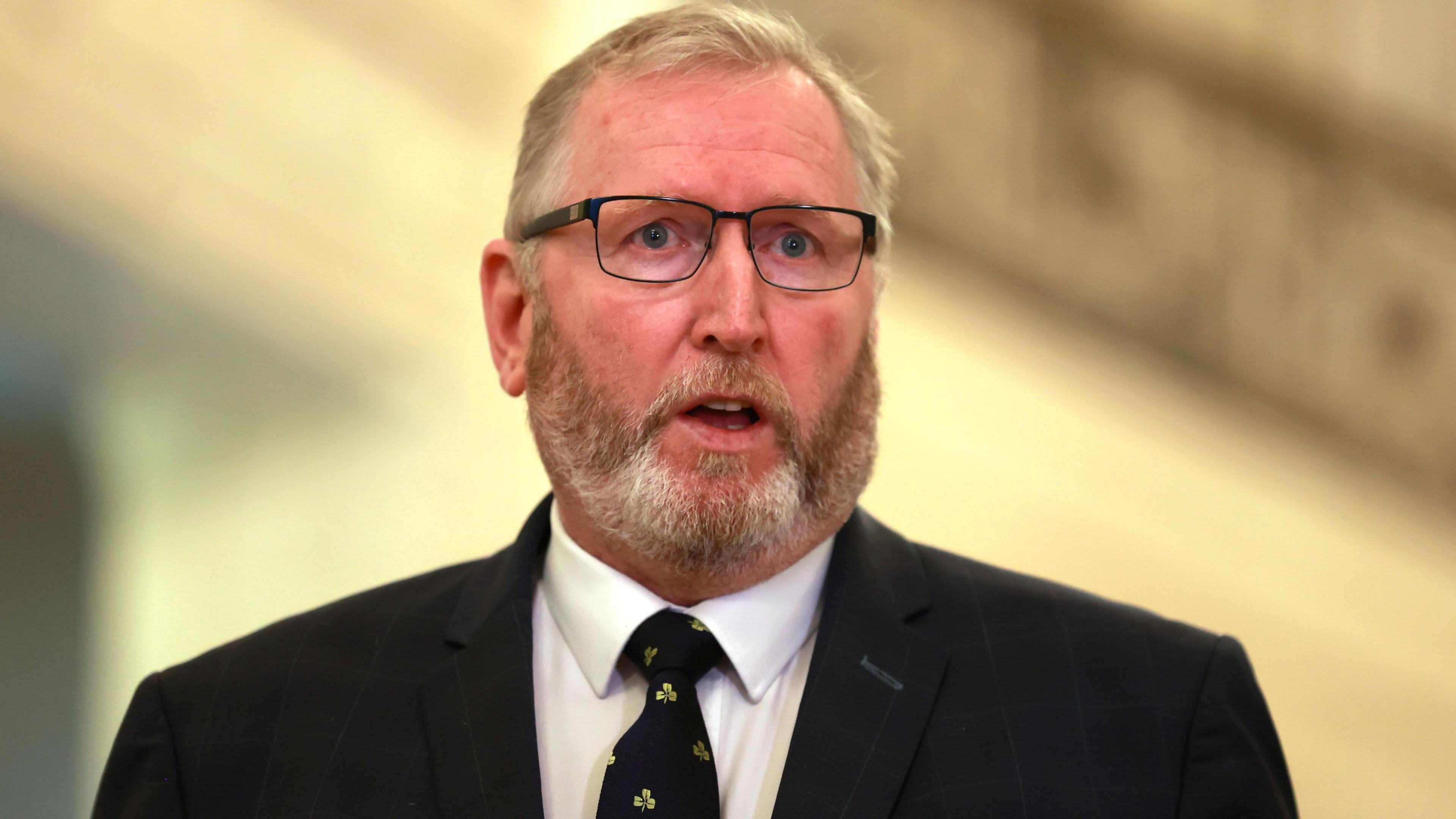 Beattie is talking in a hallway with a grand staircase. The background is blurred. He wears glasses and has a black suit on. There is a shamrock pattern on his tie. 