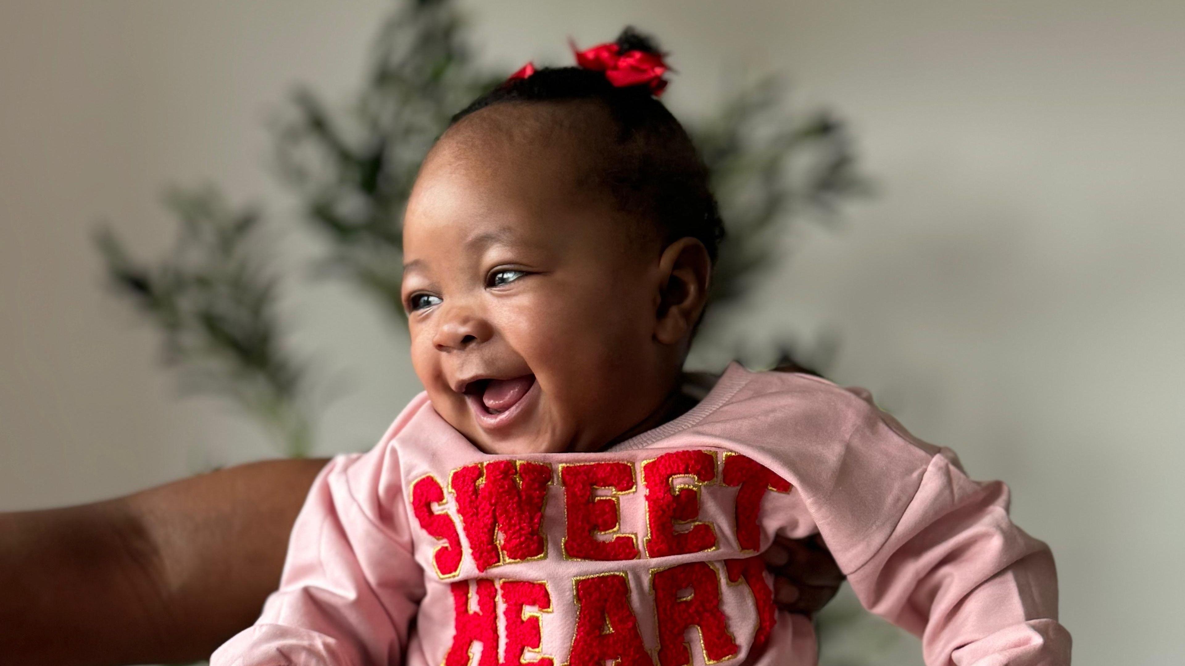A baby wearing a red hair tie and a pink jumper, which has the words "sweet heart" written on it in red. She is being held up and is looking to the left. 