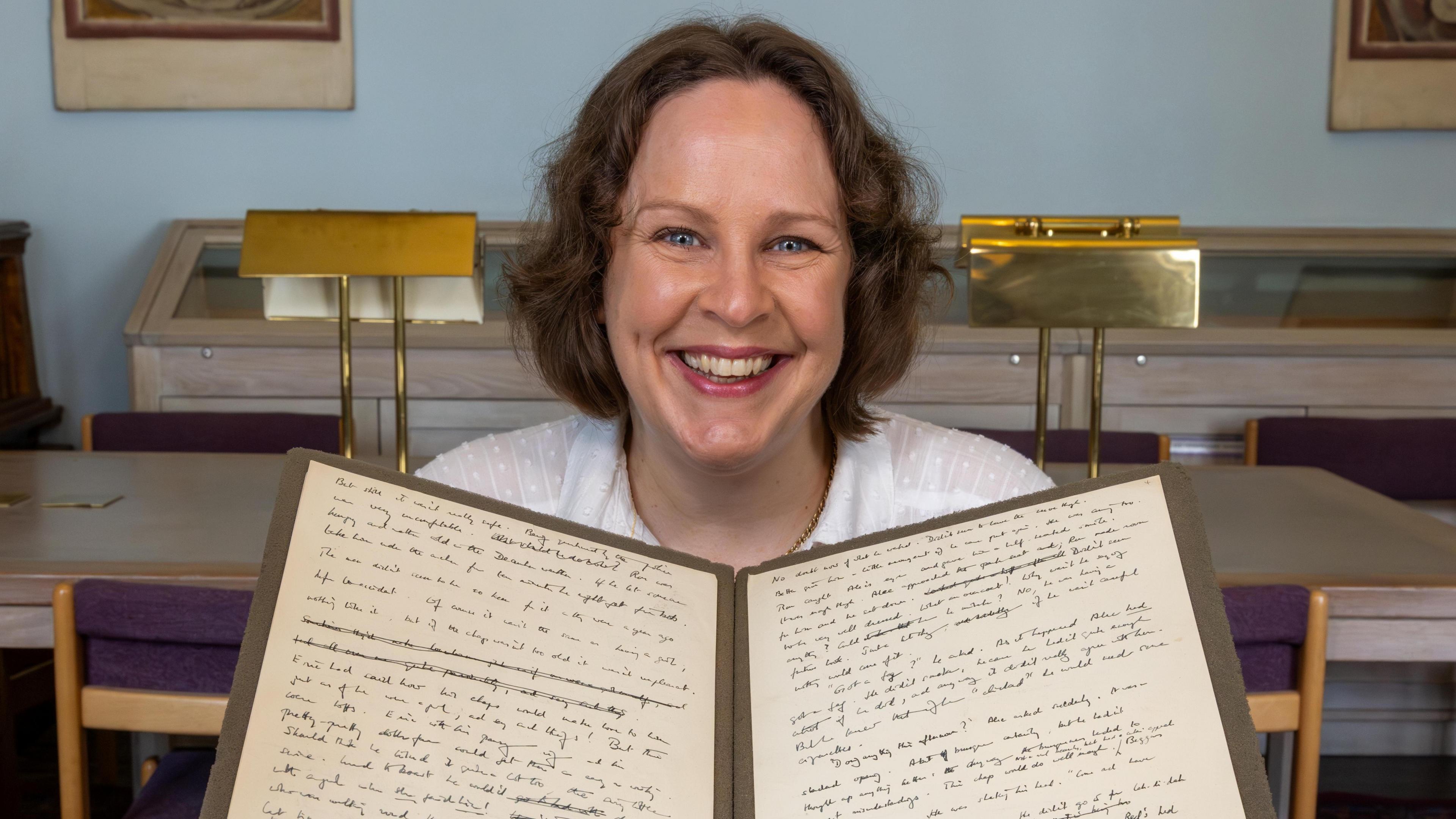 Sarah Dillon is sitting on a chair and is holding a folder which is open and has two pieces of paper with writing on it. Dillon has short brown hair and is wearing a white T-shirt.
