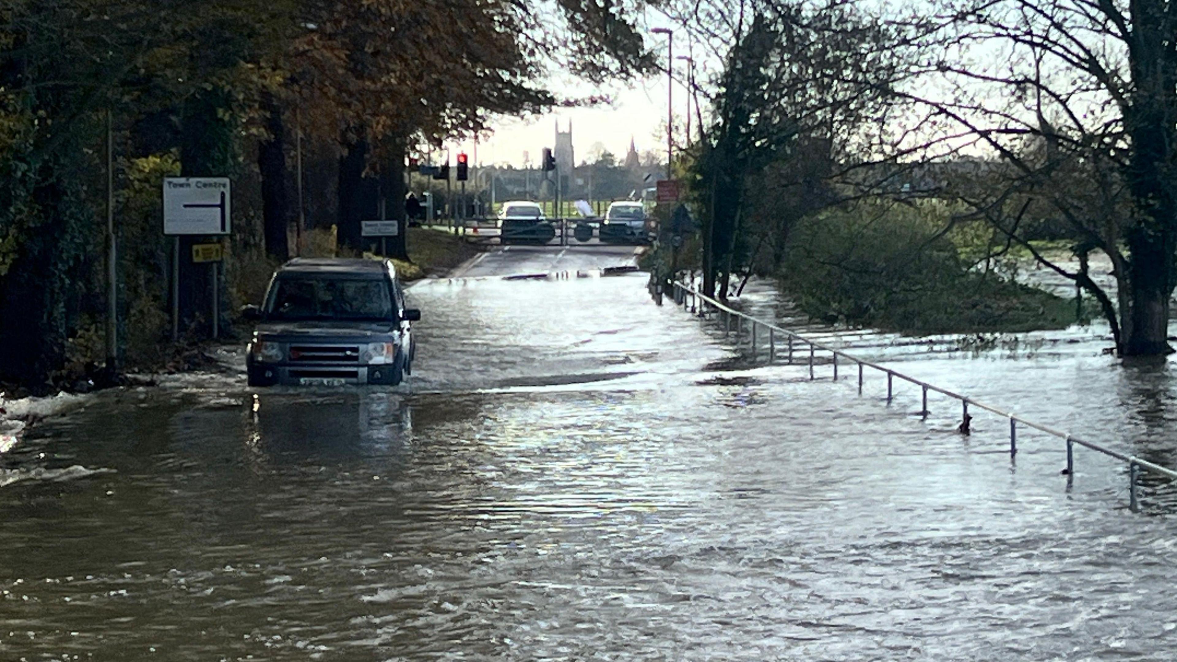 A road is submerged in water and a Range Rover is driving through, there is a sign pointing to the town centre and a church spire in the distance.