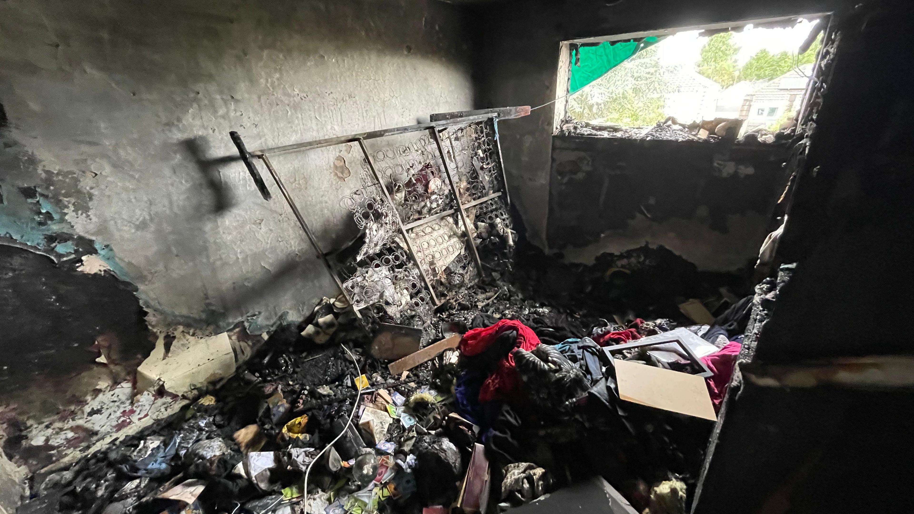 The aftermath of a fire in a small room. The room is severely damaged, the walls are blackened and soot-covered, and the floor is littered with the remains of furniture, personal belongings, and debris