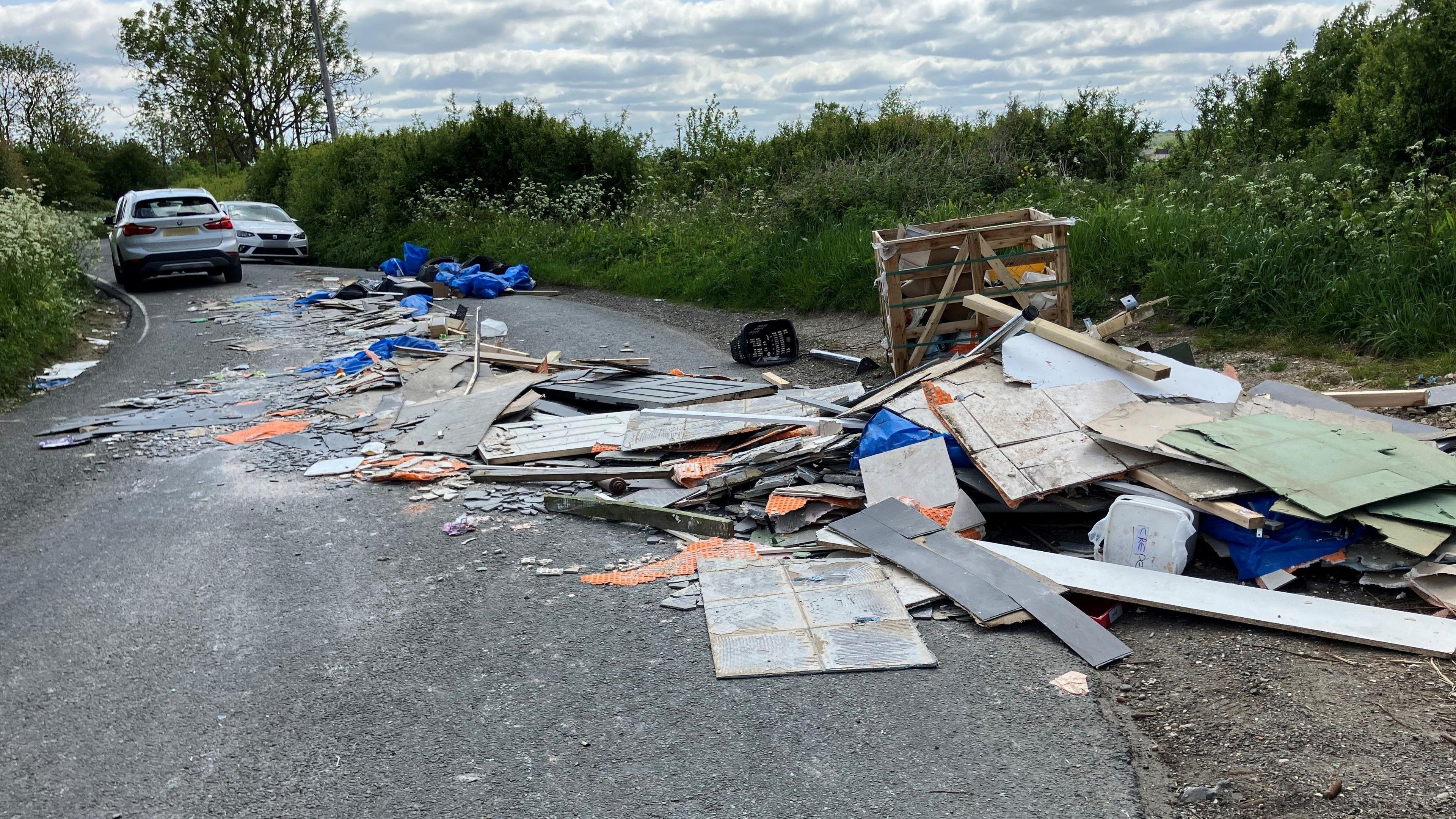 A large amount of litter spread out across the bend on a country lane. Cars are waiting to fit through the narrow gap.