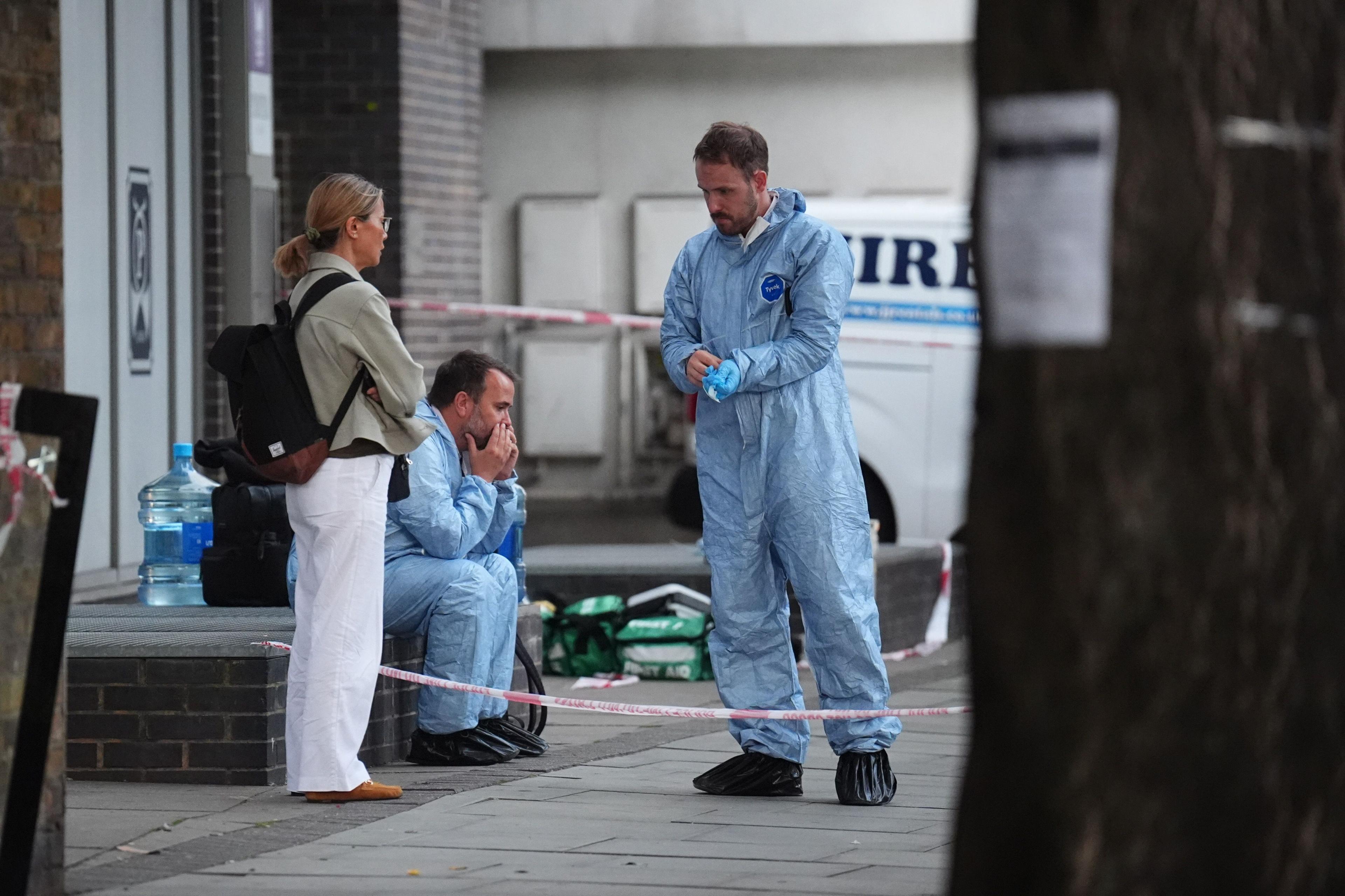 Two police officers in blue forensic overalls behind police tape speak to a woman in white trousers who is wearing a black rucksack and folding her arms