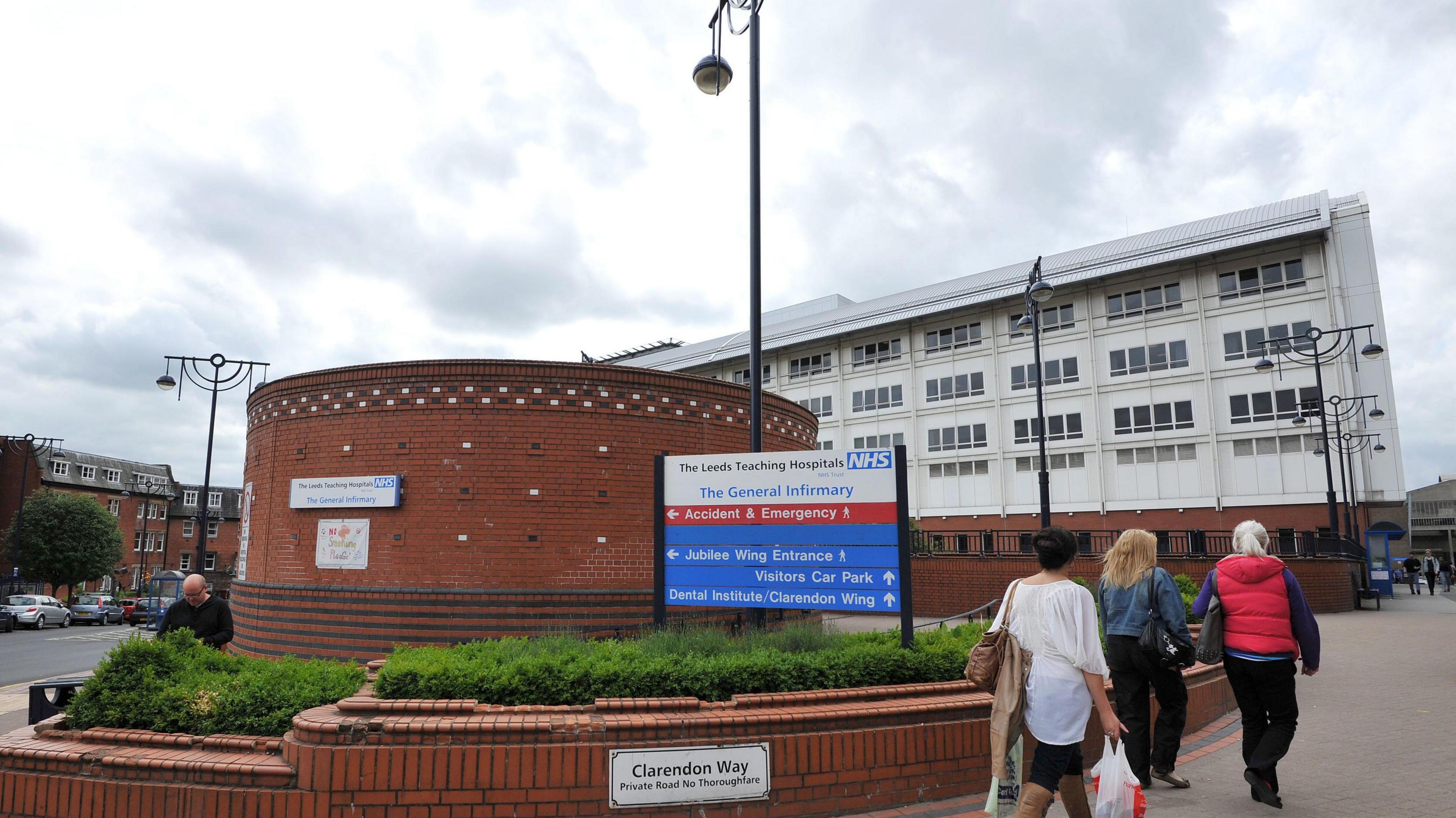 Main entrance to Leeds General Infirmary - with a circular brick building with a large sign reading Leeds Teaching Hospitals NHS Trust and directions to parts of the site. There is a large white building behind.
