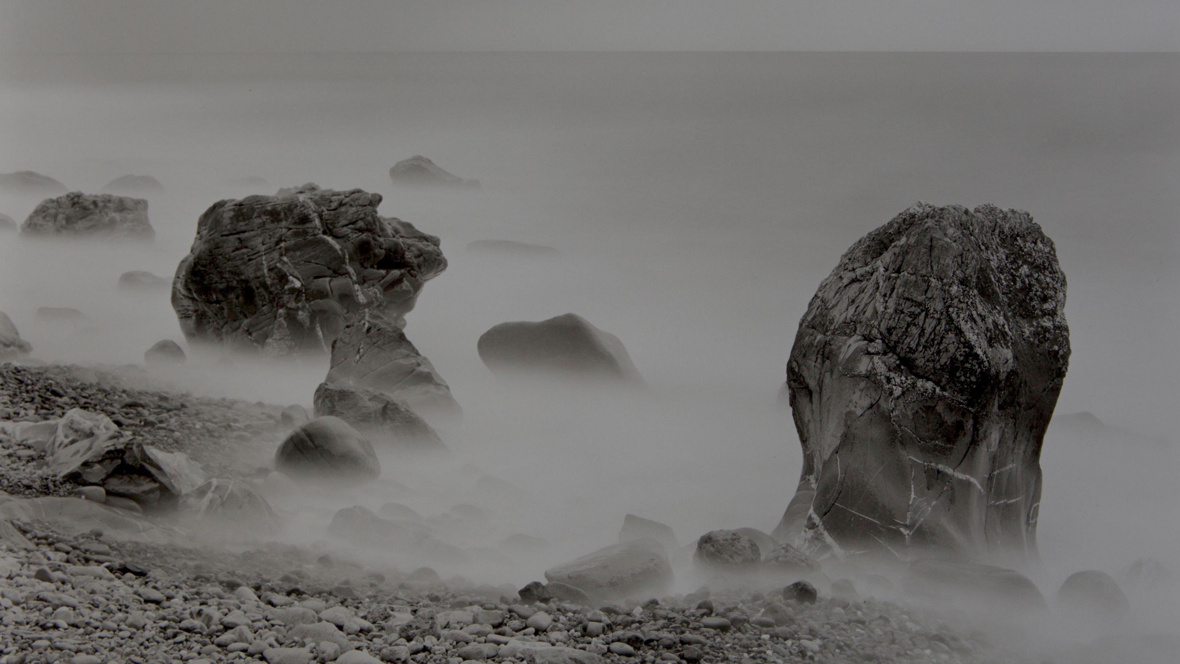 A work by John Blakemore featuring mist surrounding larger stones on a pebble beach