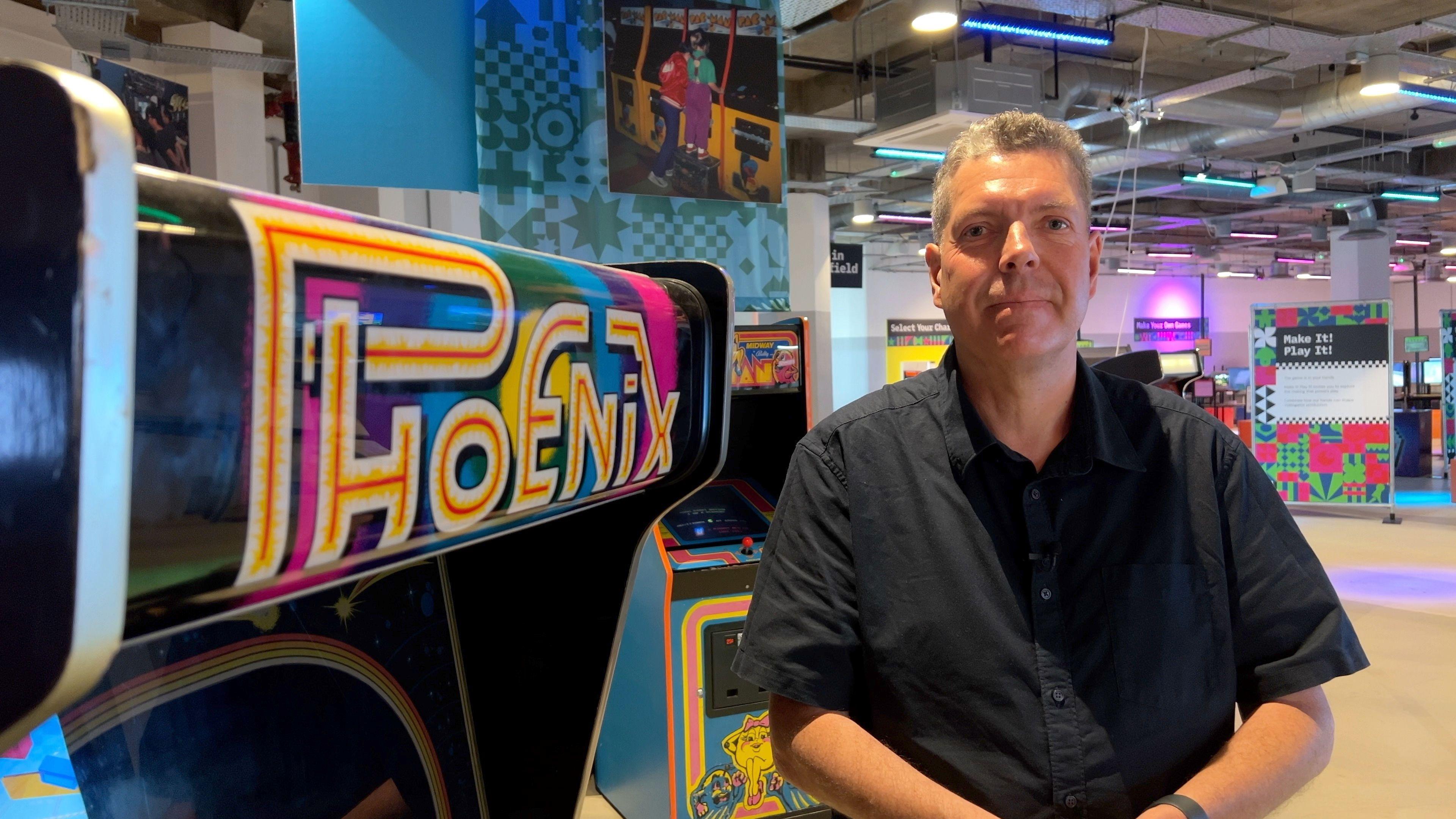 Jamie Sefton from gaming industry body Game Republic wears a darks shirt with his hands clasped in front of him. He has grey hair and is smiling. He is standing in front of the Phoenix arcade machine at the National Video Games Museum in Sheffield
