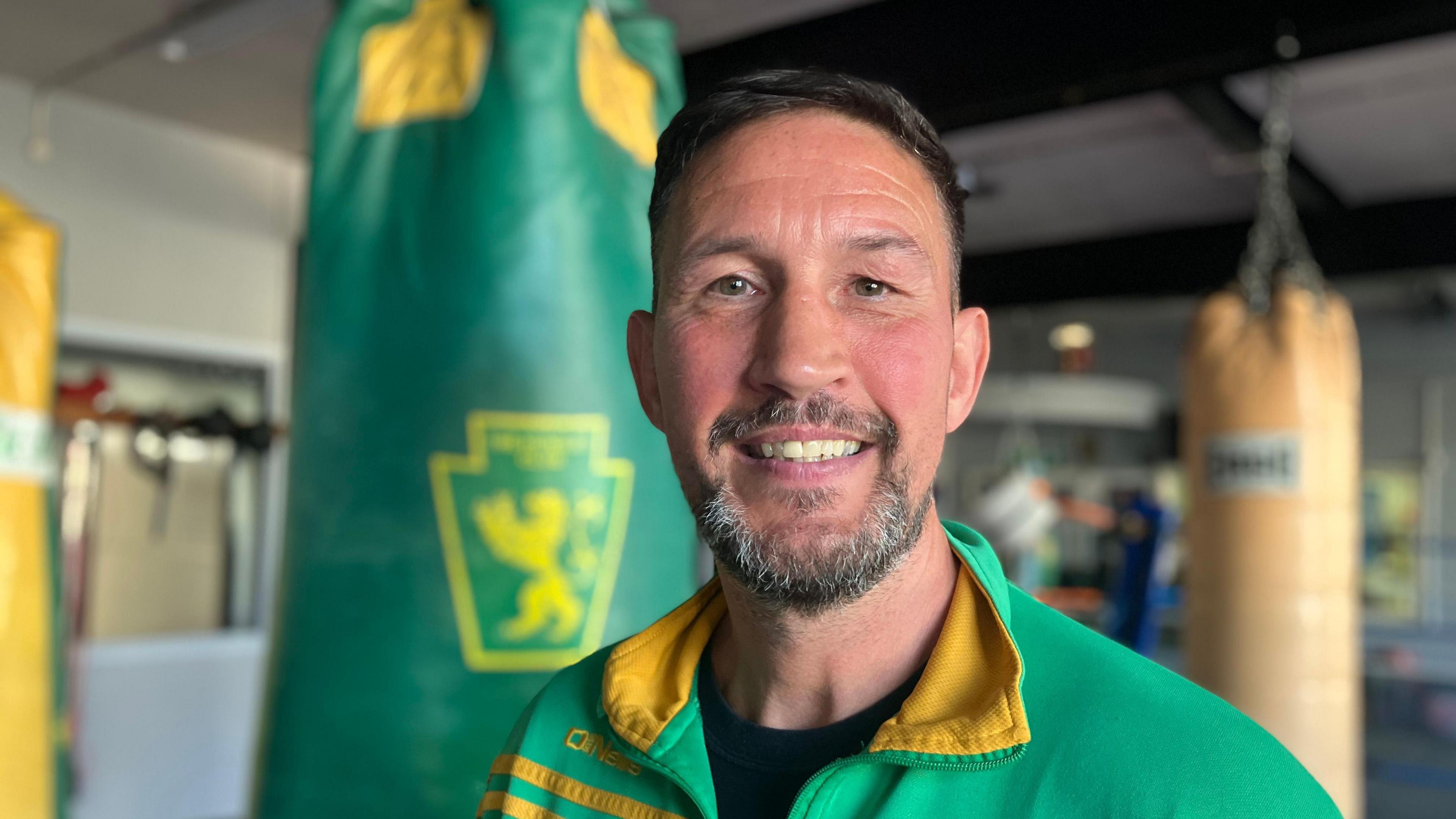 A man with dark hair and a beard smiles at the camera. He wears a green tracksuit with a yellow trim. Behind him, hang green and yellow punchbags.