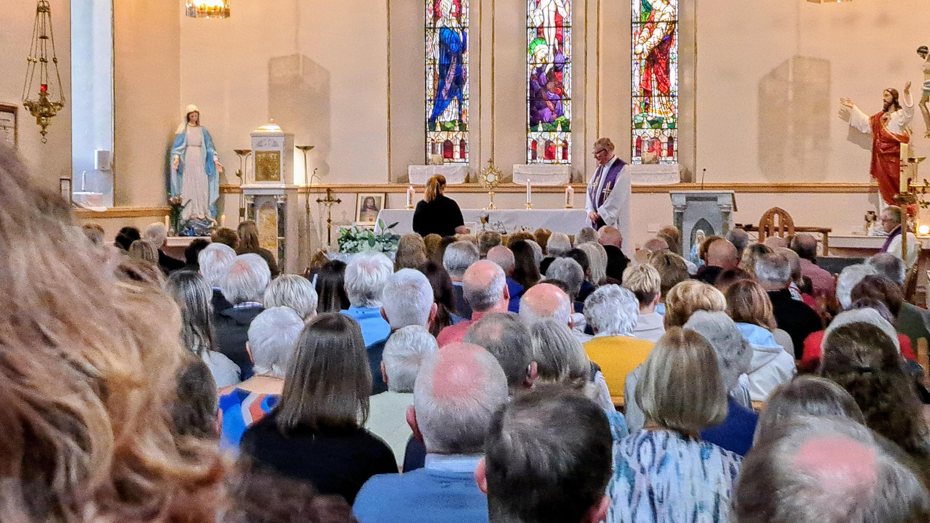 The interior of a church. A service is being held. The congregation is sitting. A priest stands at a the altar.