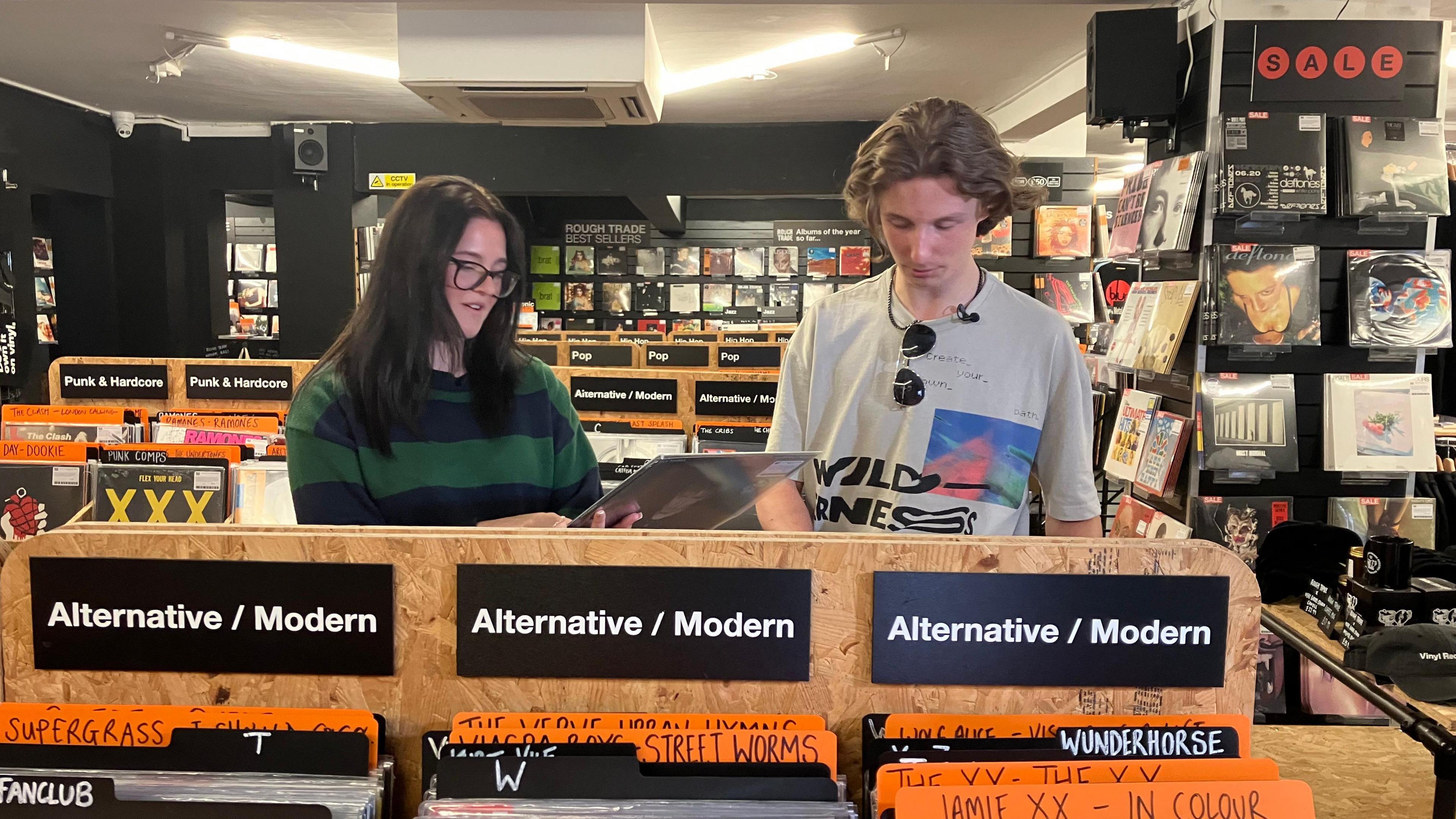 Hermione and Max browse records in a shop. The names of music genres plus acts like Supergrass and Jamie XX can be seen on cards above the records.