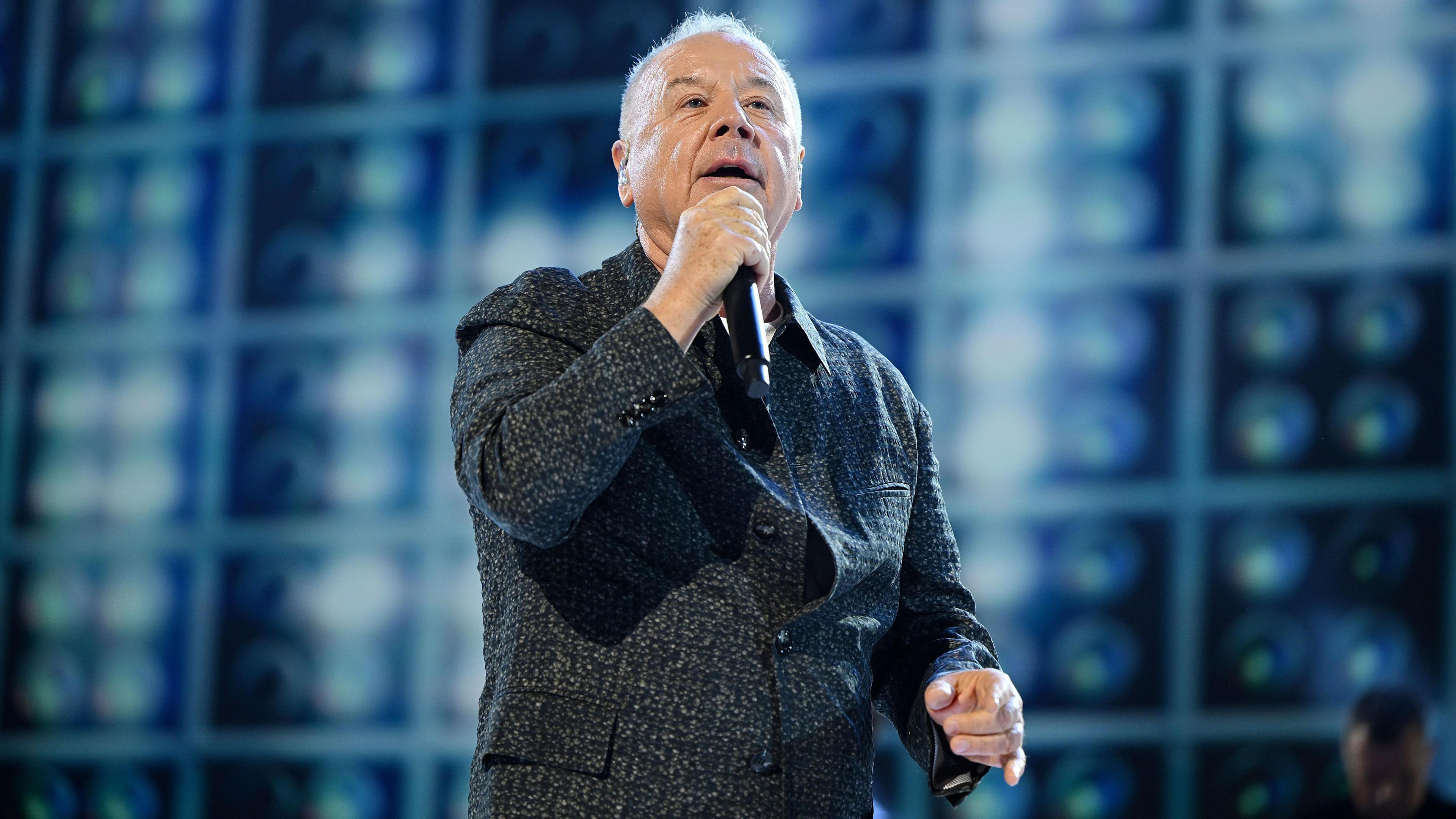 Jim Kerr of Simple Minds in a grey jacket on stage singing. He is in front of a blue textured wall.