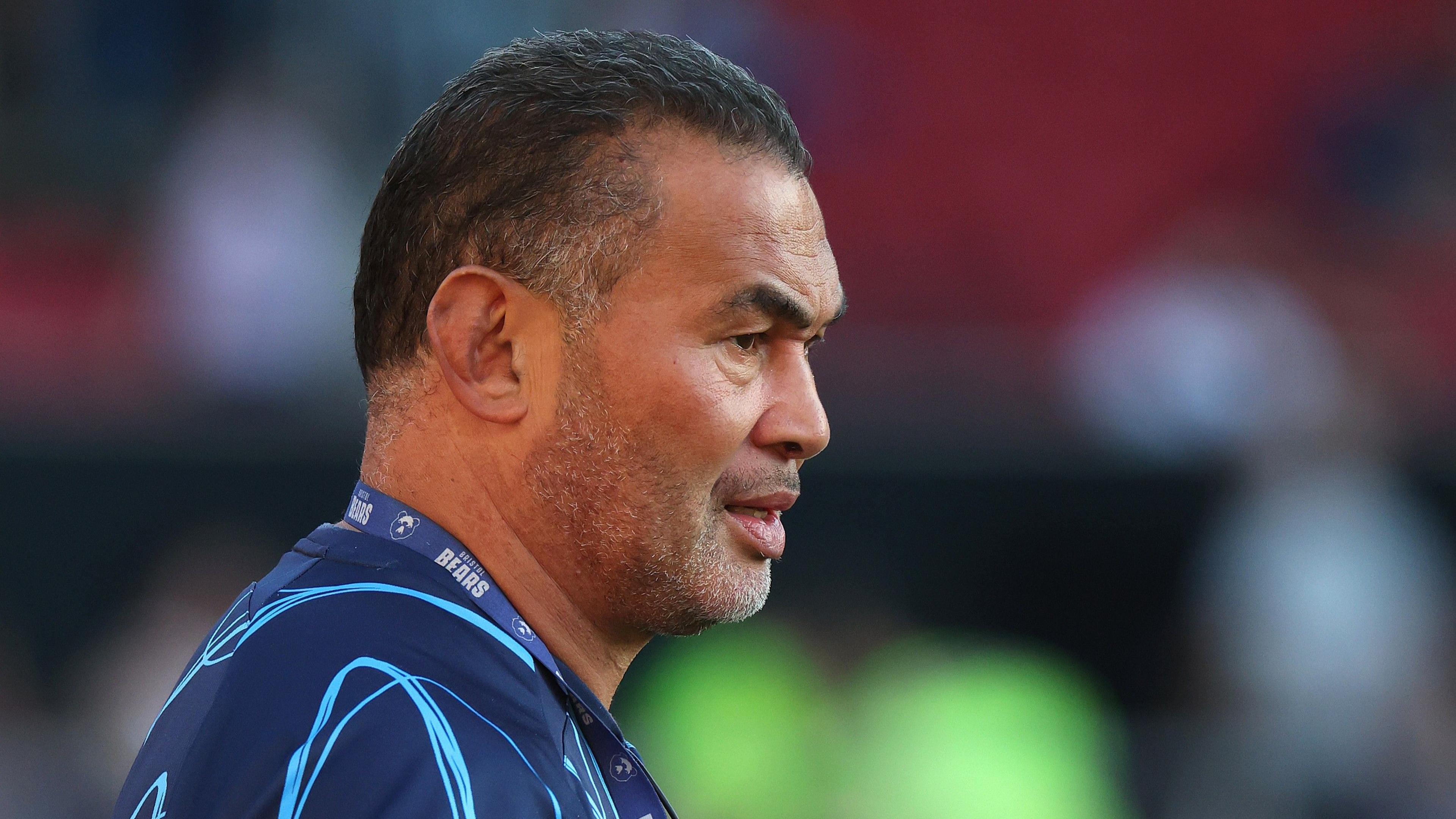 Side profile image of Bristol Bears director of rugby Pat Lam wearing a blue shirt and a lanyard with a blurred background.