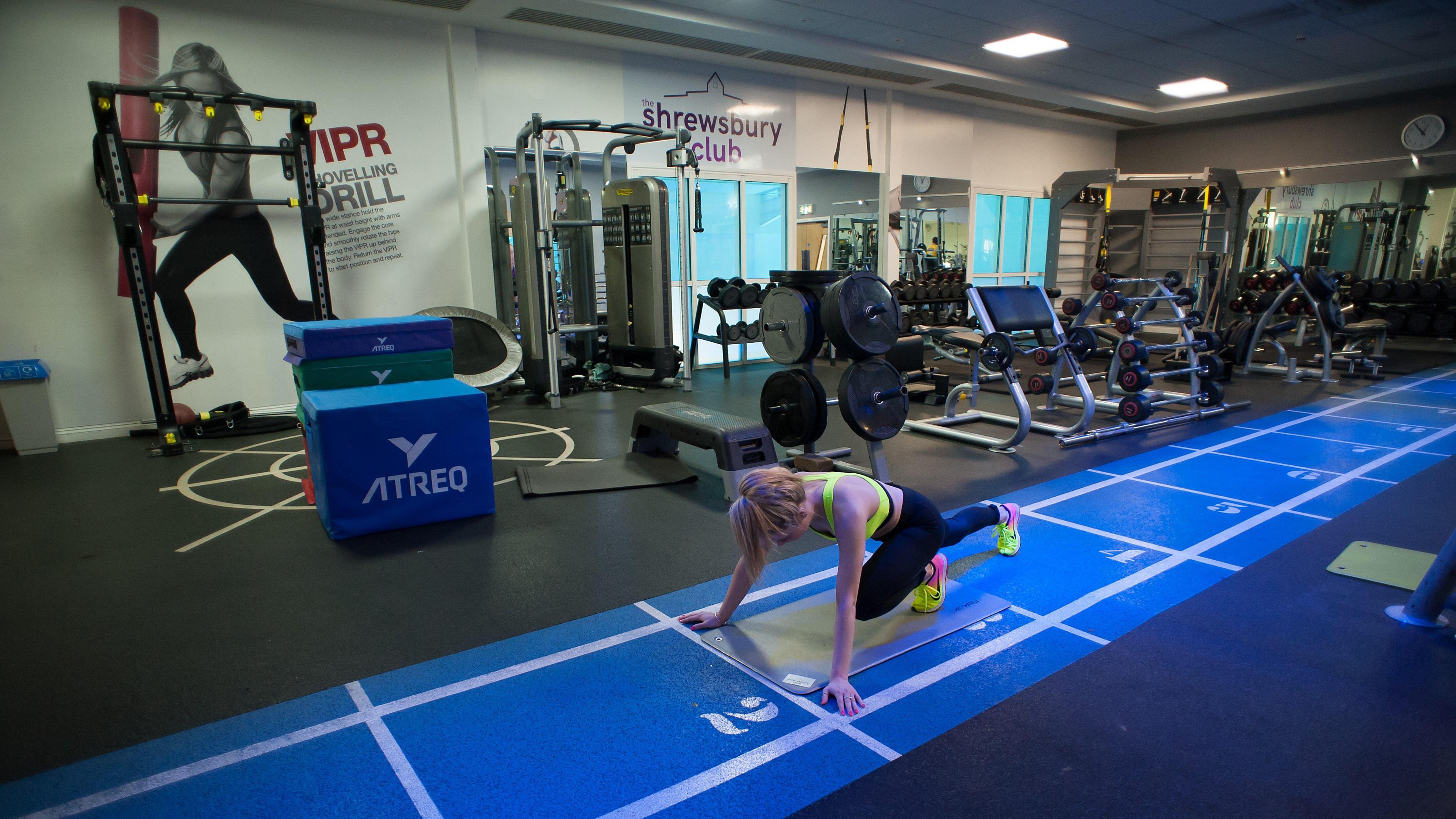 An indoor gym with lots of equipment. There is a blue track on the floor, with a woman exercising on it.