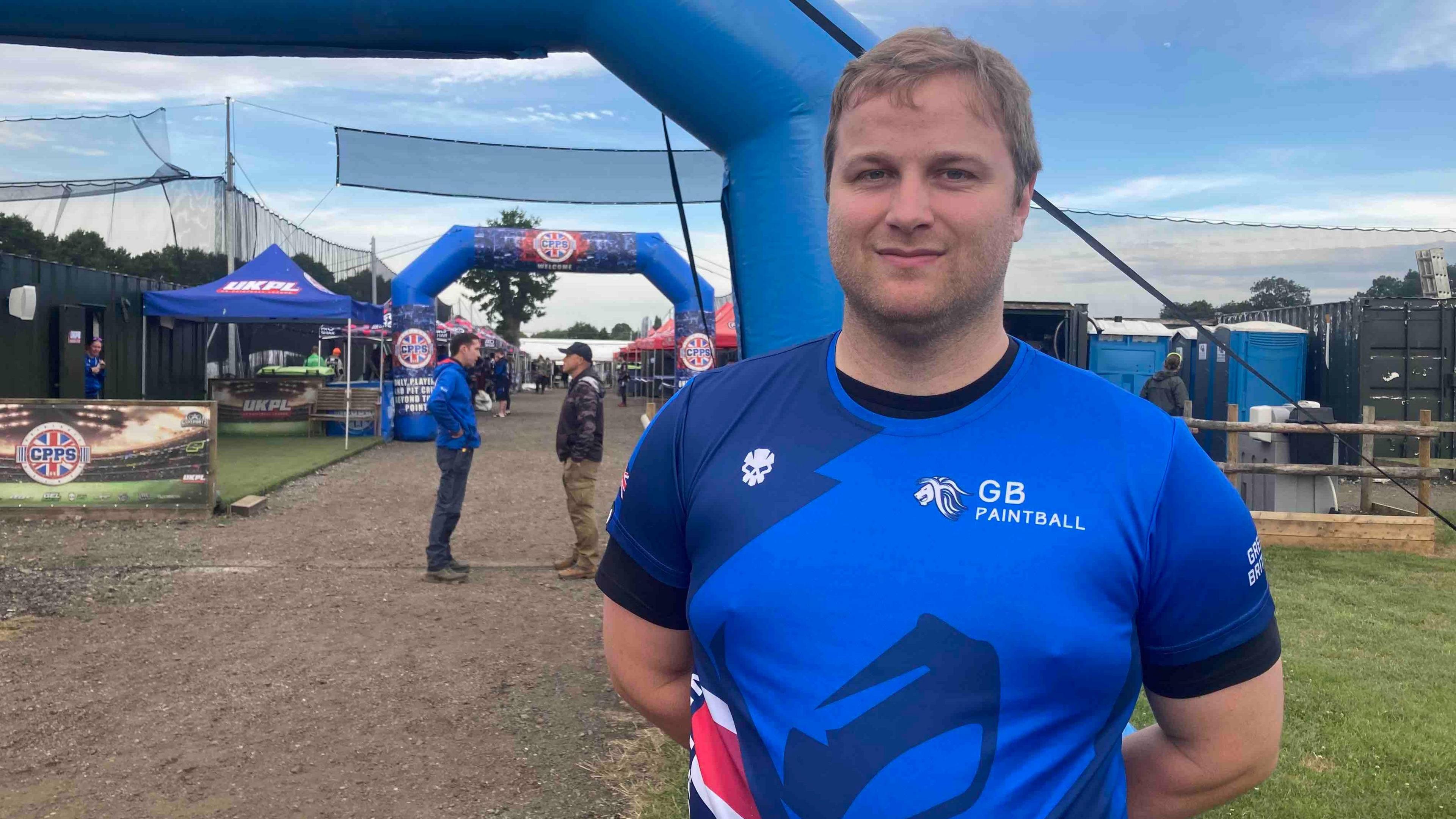 A man stands outside. He has short blonde hair and he is wearing a blue sports T-shirt with "GB Paintball" on it in white letters.