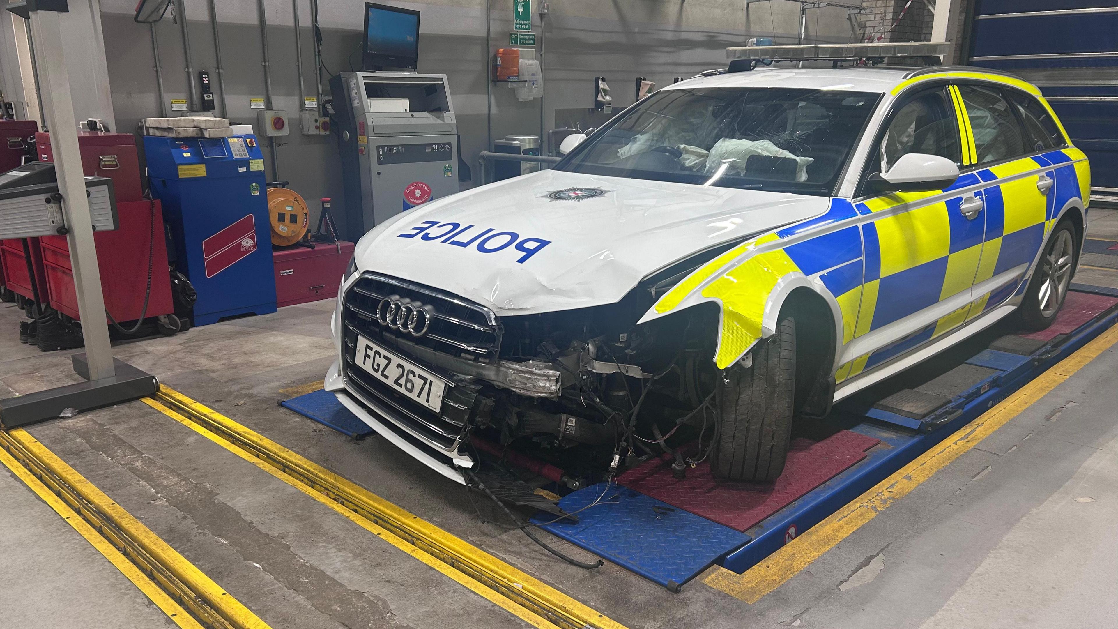 A white police Audi has yellow and blue patterns on its sides. Their is a large dent and missing panel from the front left of the car, on the passegers side