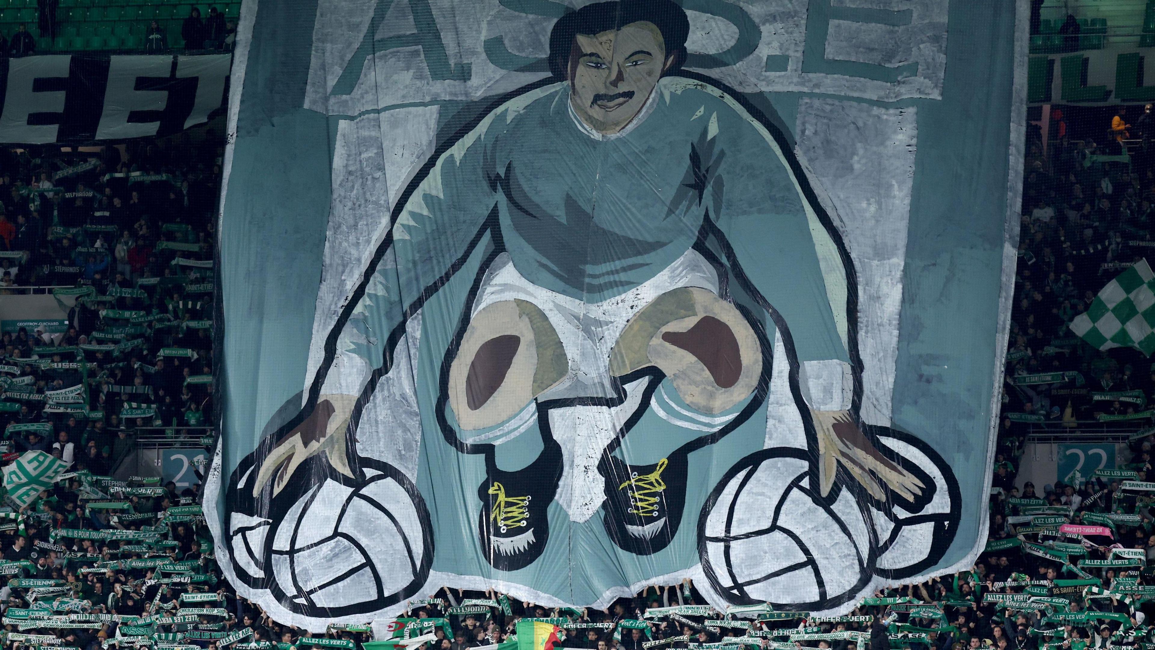 A cartoon-like large green and white drawing of Rachid Mekhloufi crouching down with each hand on two footballs is raised by football supporters in a stand, while others hold green and white St-Etienne scarves aloft with outstretched arms