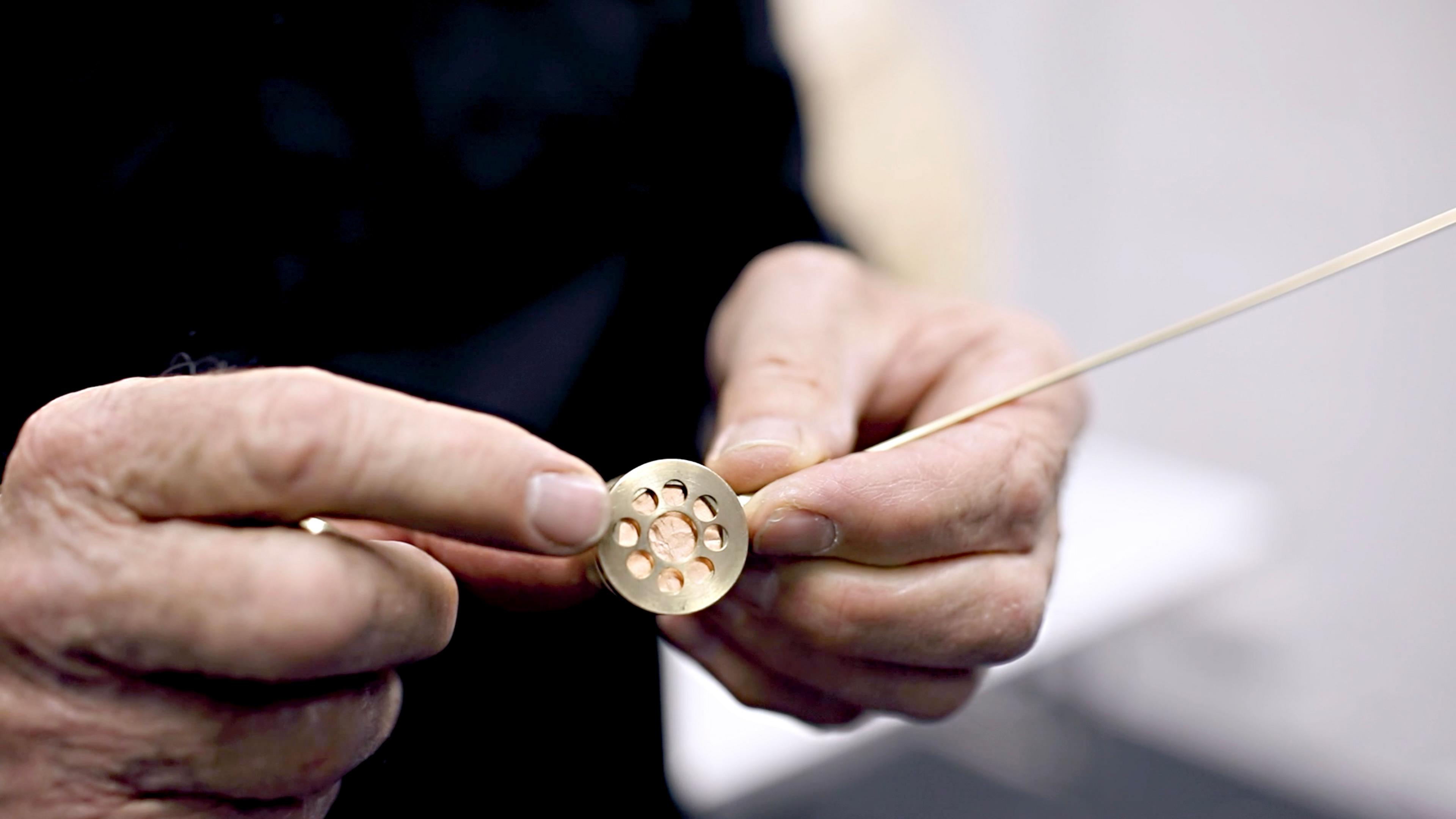 A close of The Thing, with a man's hand holding it, it is a gold circle with holes cut out, with a long gold stick coming out of it.