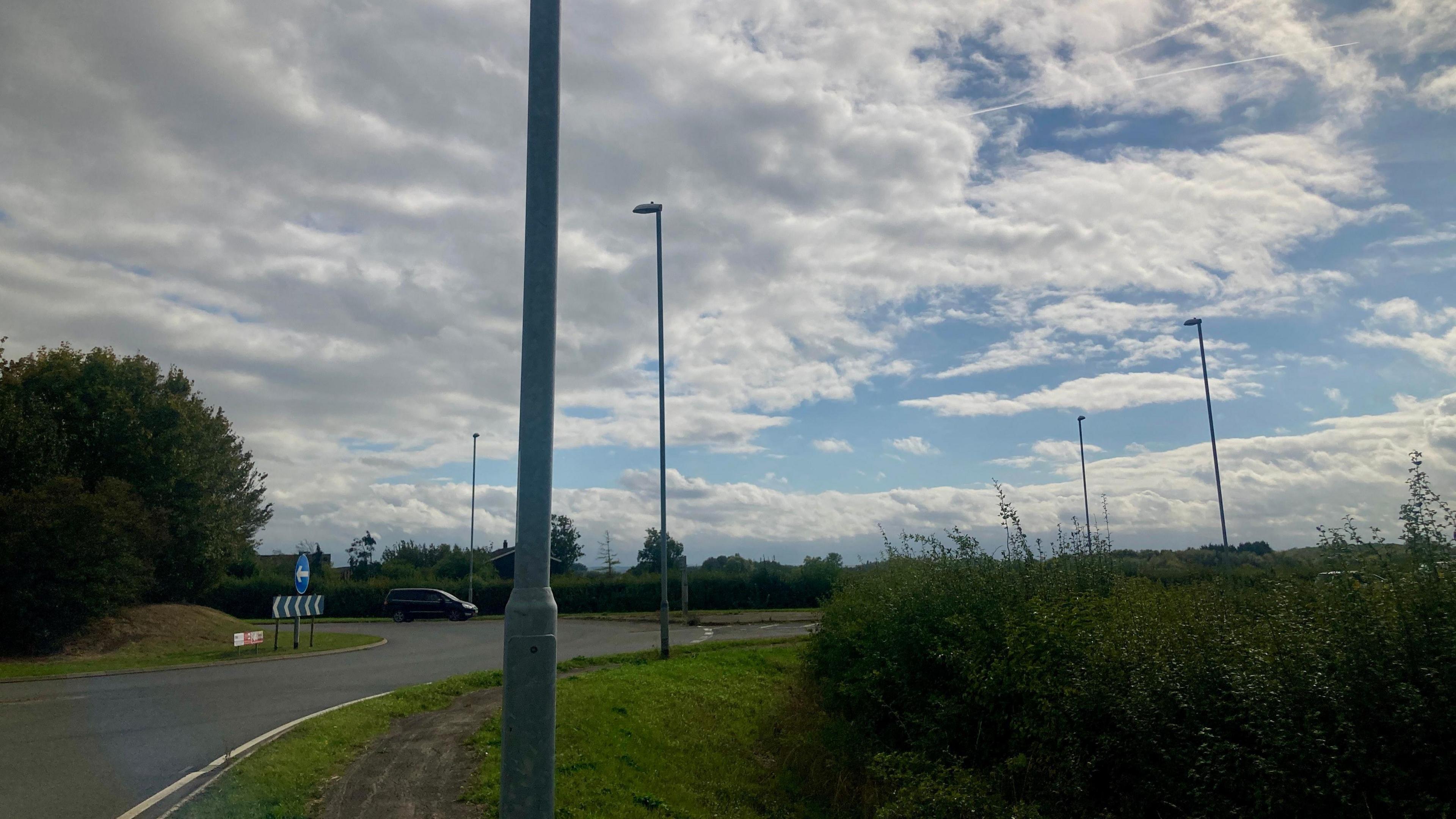 Another view of a roundabout. Lampposts can be seen on the edge of the roundabout. A van can be seen exiting the roundabout in the distance. 