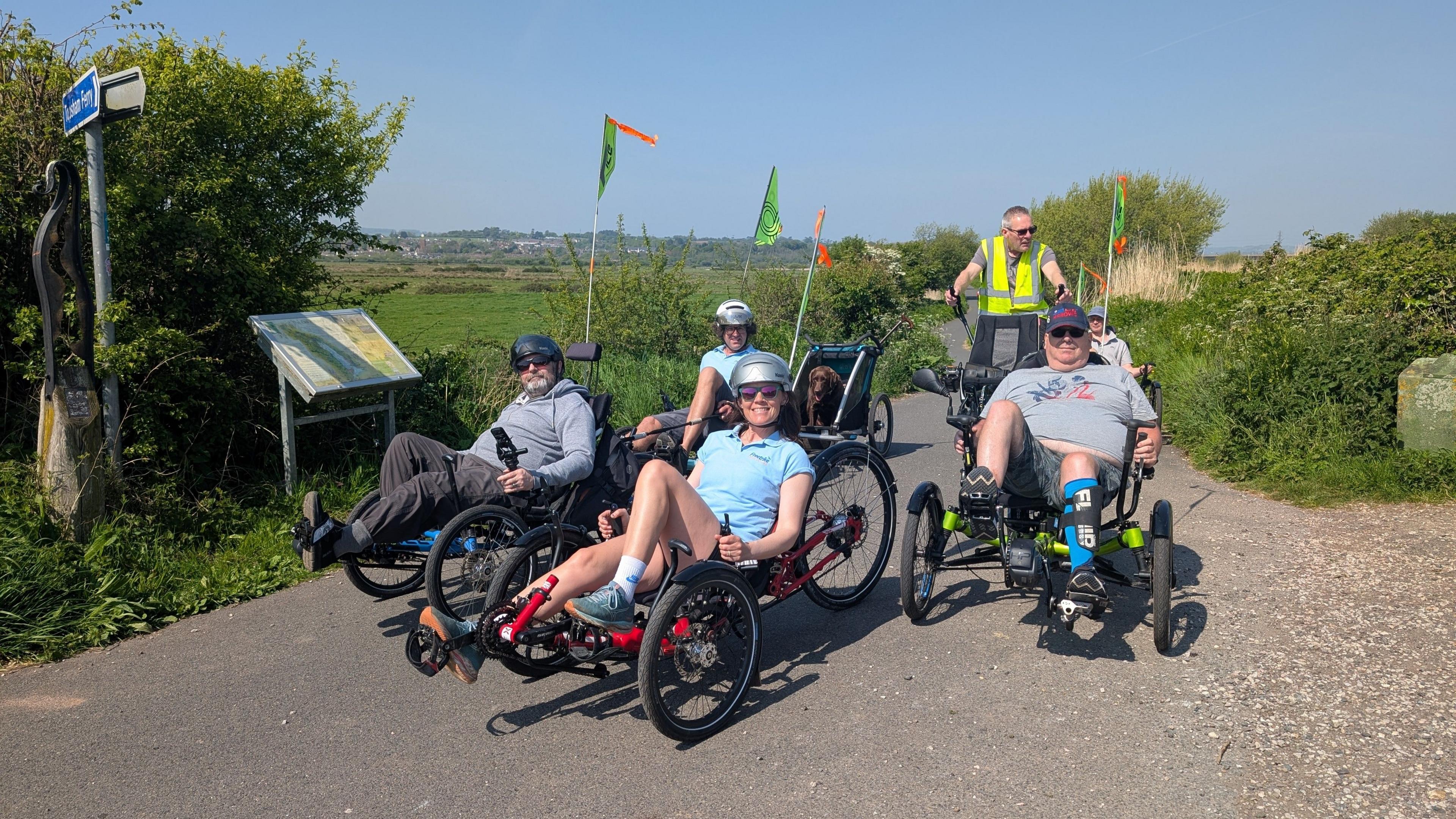Four people on recumbent trikes smile for the camera. They are out on the Exe Estuary Trail on a sunny day.