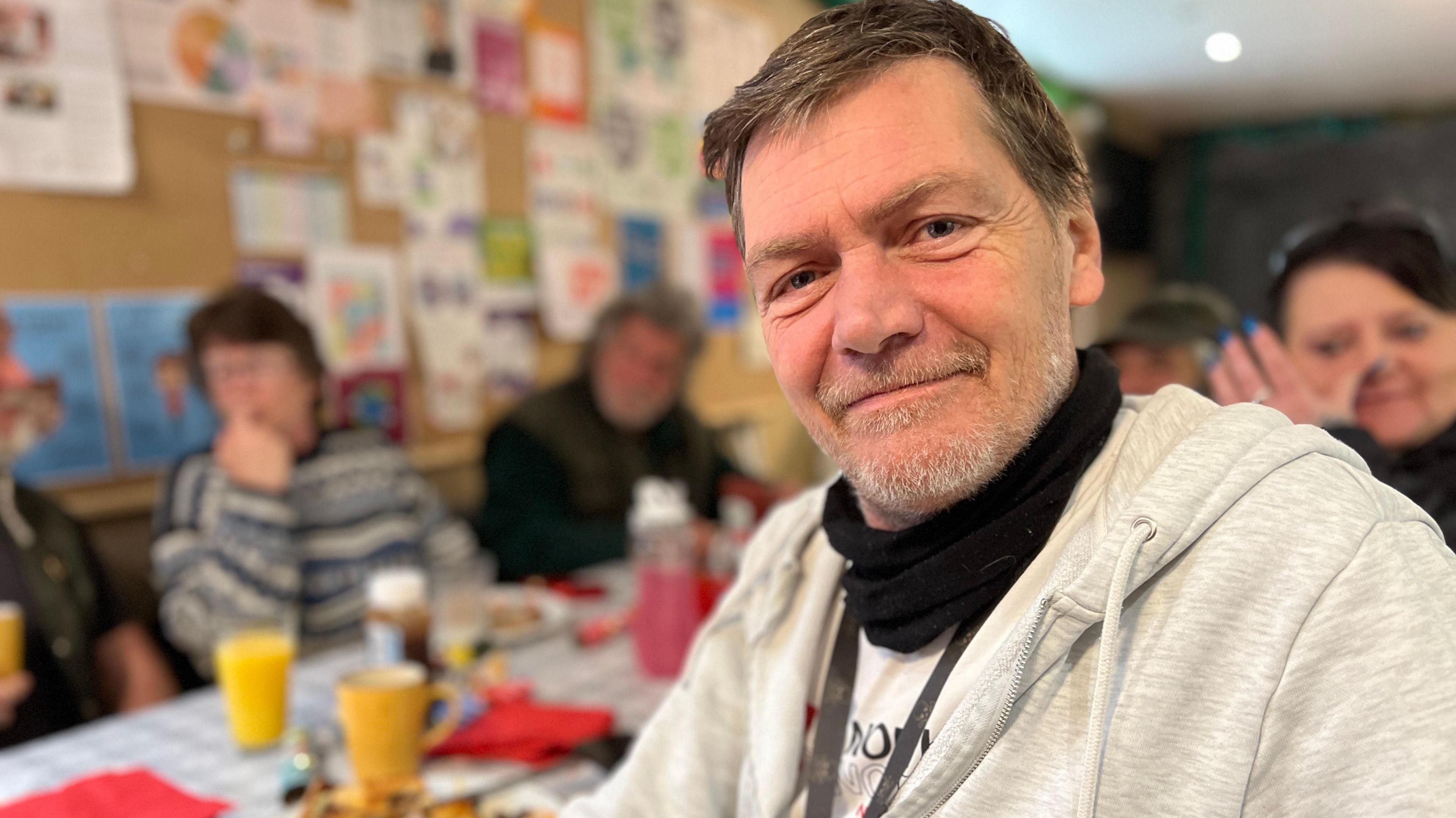 Alistair at a table containing mugs and plates of food. He is wearing a light grey zip top and a black scarf and is smiling at the camera.