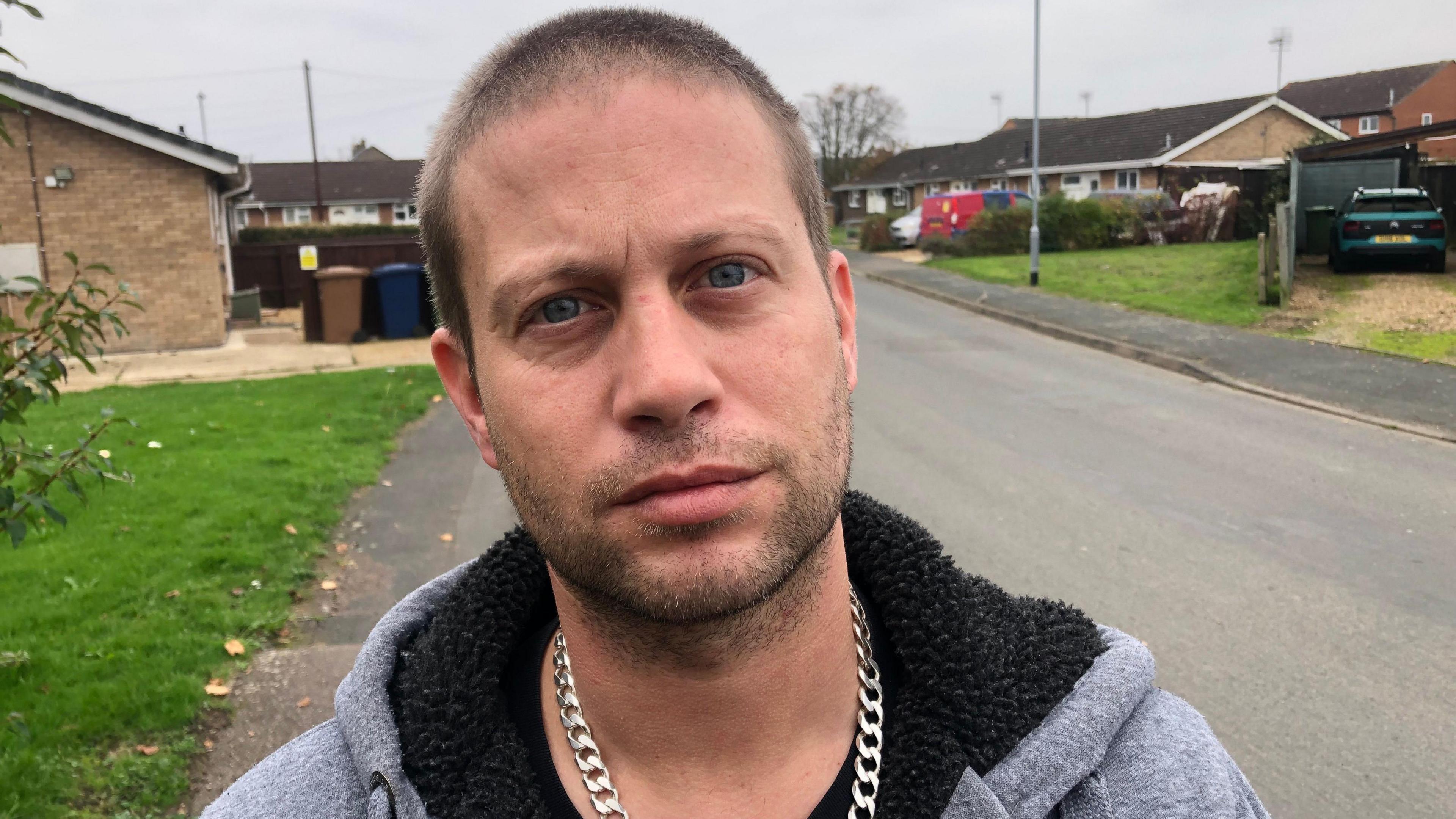 Lee Darling, 37, has a grey fleece with black lining in the hood, he had a thick silver chain around his neck. He has stubbly chin and short dark cropped hair, behind him is Papworth Road, where the attack took place, bungalows either side of the street.