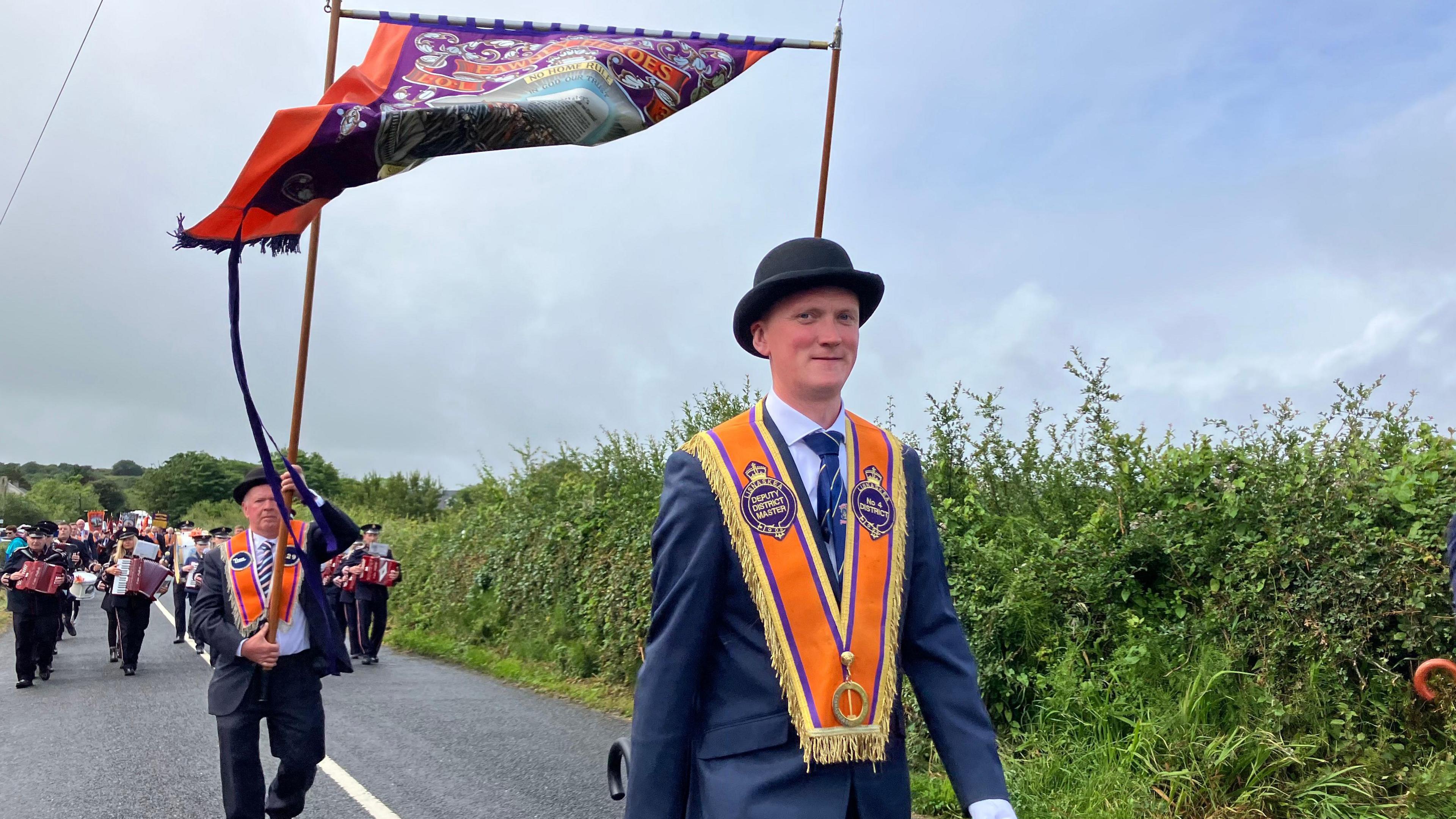 A man in a blue suit has an orange sash around his shoulders and a black bowler hat. There is a group of marchers following behind with an array of instruments