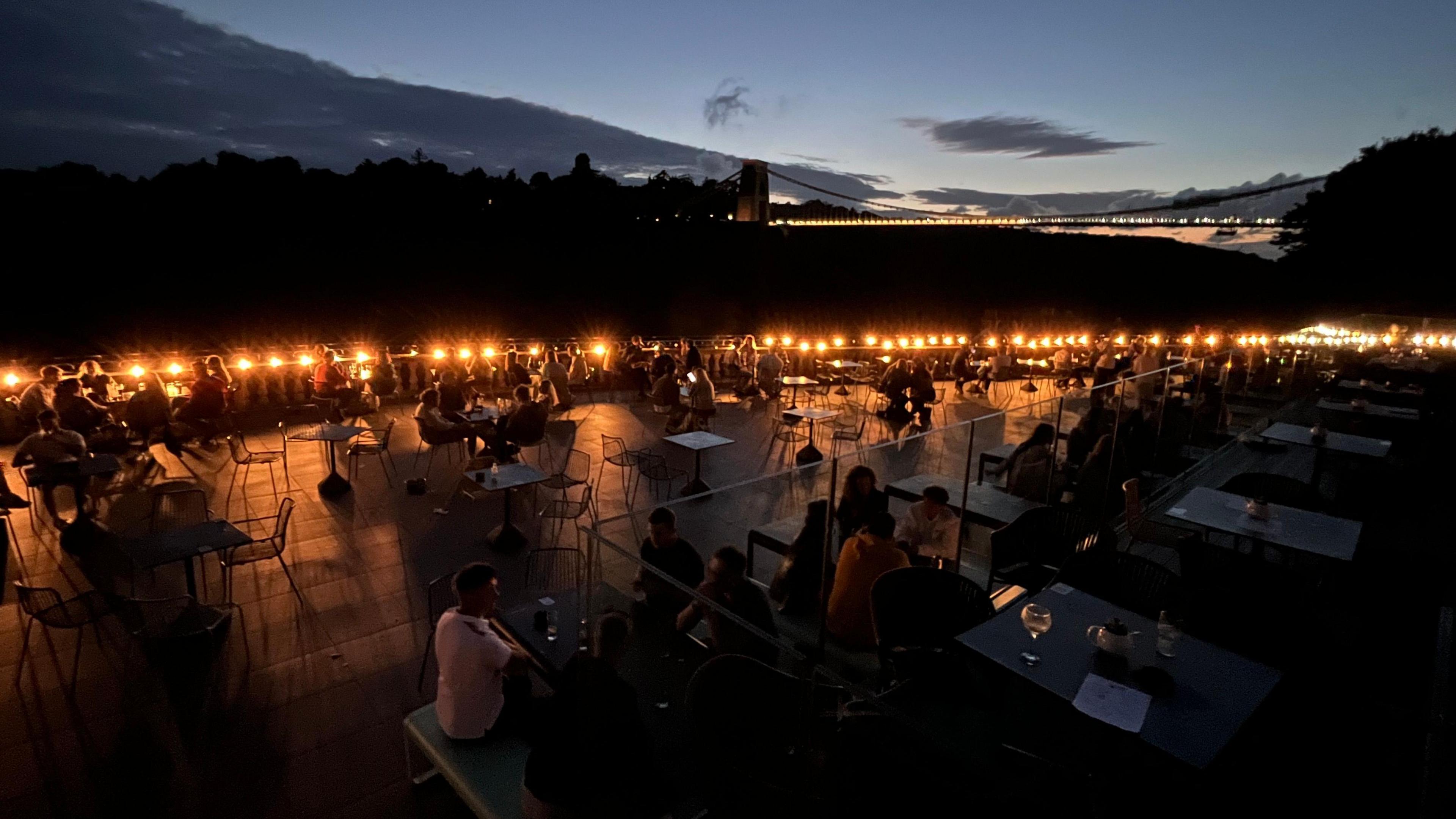 People sit in the glow of lamps at the White Lion Bar in Clifton in Bristol. In the distance the outline of the illuminated Clifton Suspension Bridge can be seen