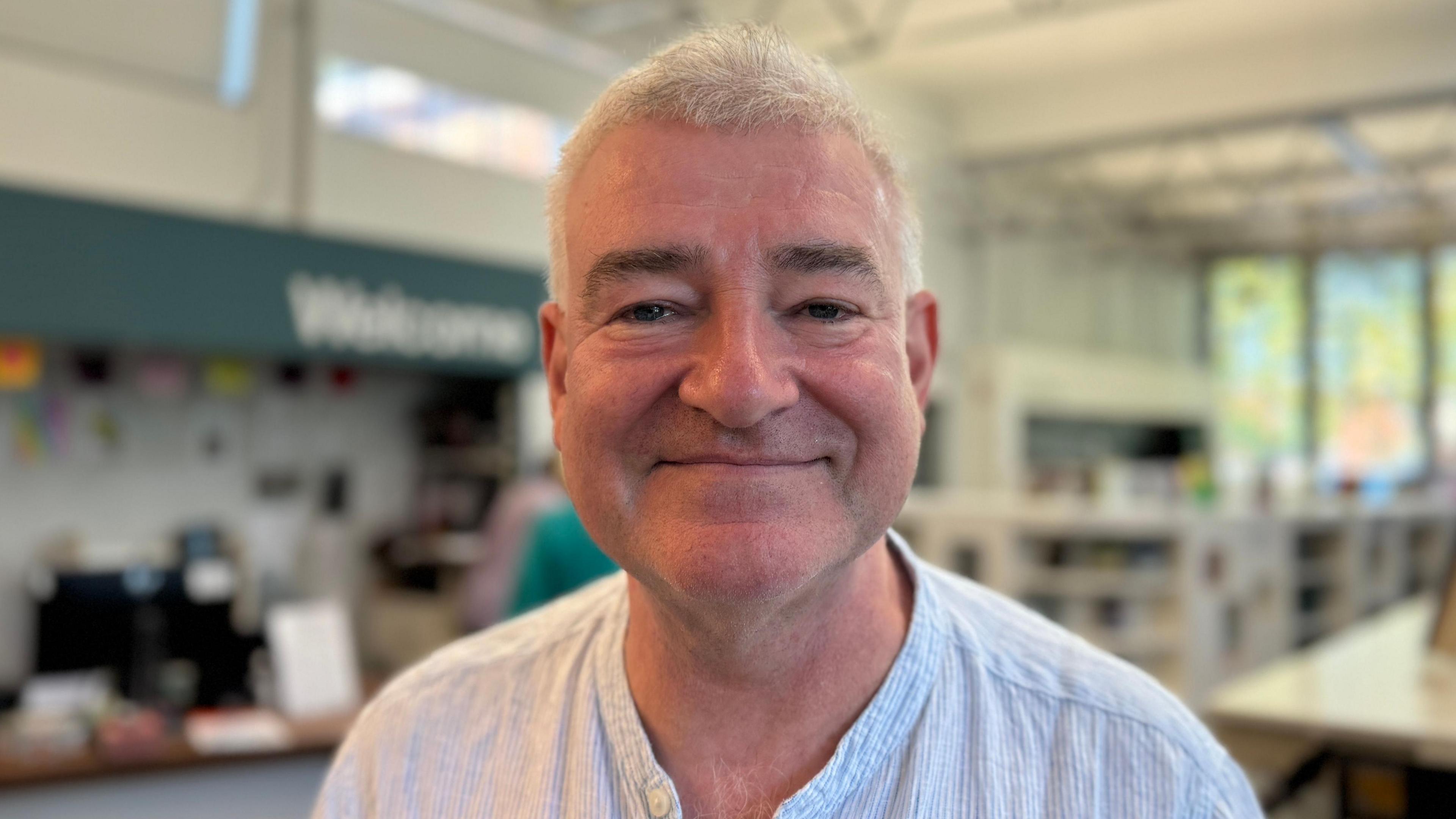 Gerry Turbitt - a man with short grey hair wearing a striped shirt smiles at the camera. He is standing in front of a blurred Welcome sign in the library.