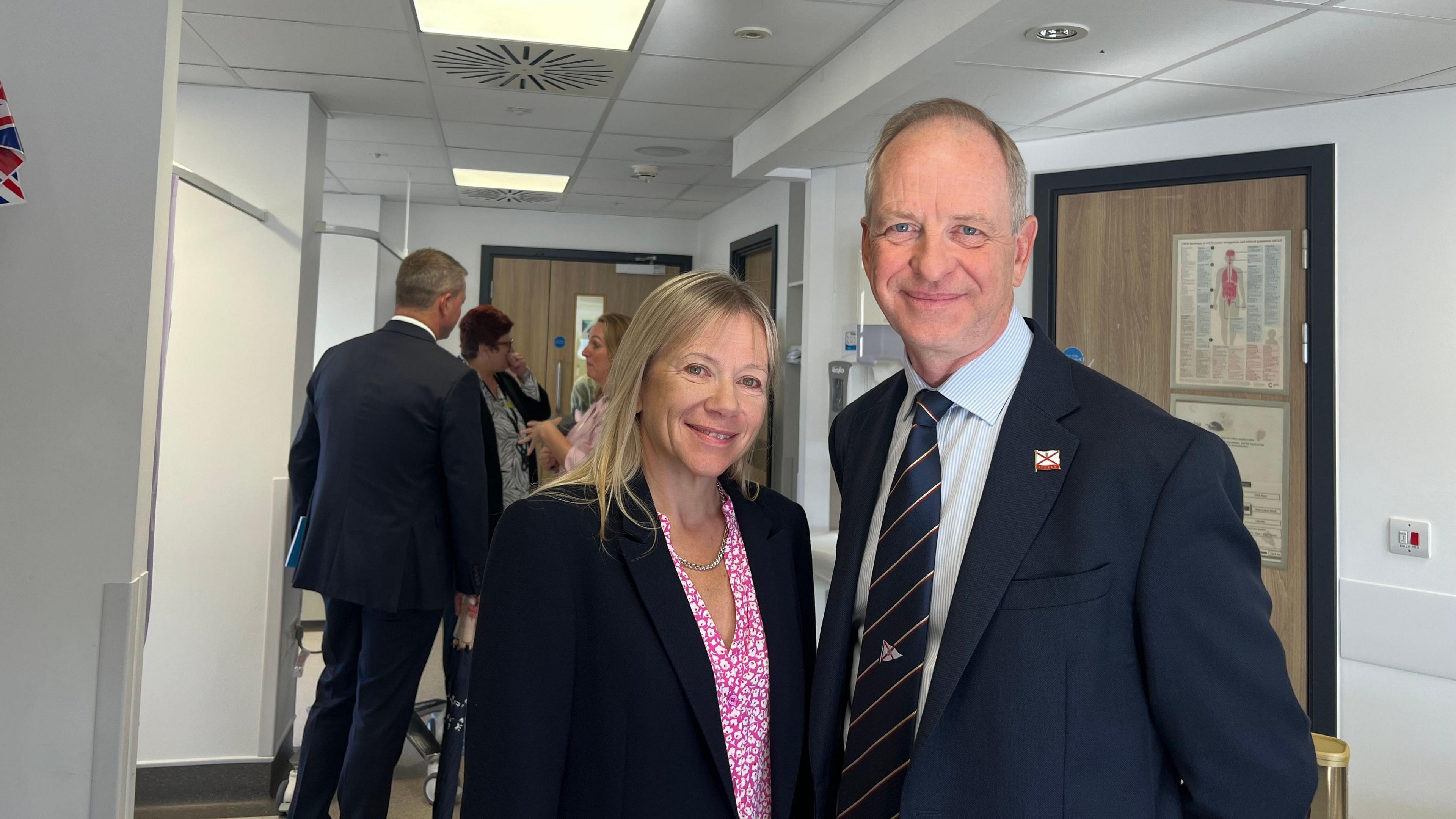 Lieutenant Governor of Jersey Vice Admiral Jeremy Kyd, with his wife Dr Karen Kyd. The Lt Gov is wearing a navy blazer, light blue shirt and a blue and red-striped tie. Mrs Kyd is wearing a black blazer and a pink and white dress. They are stood shoulder to shoulder and are both smiling at the camera in a hospital corridor.