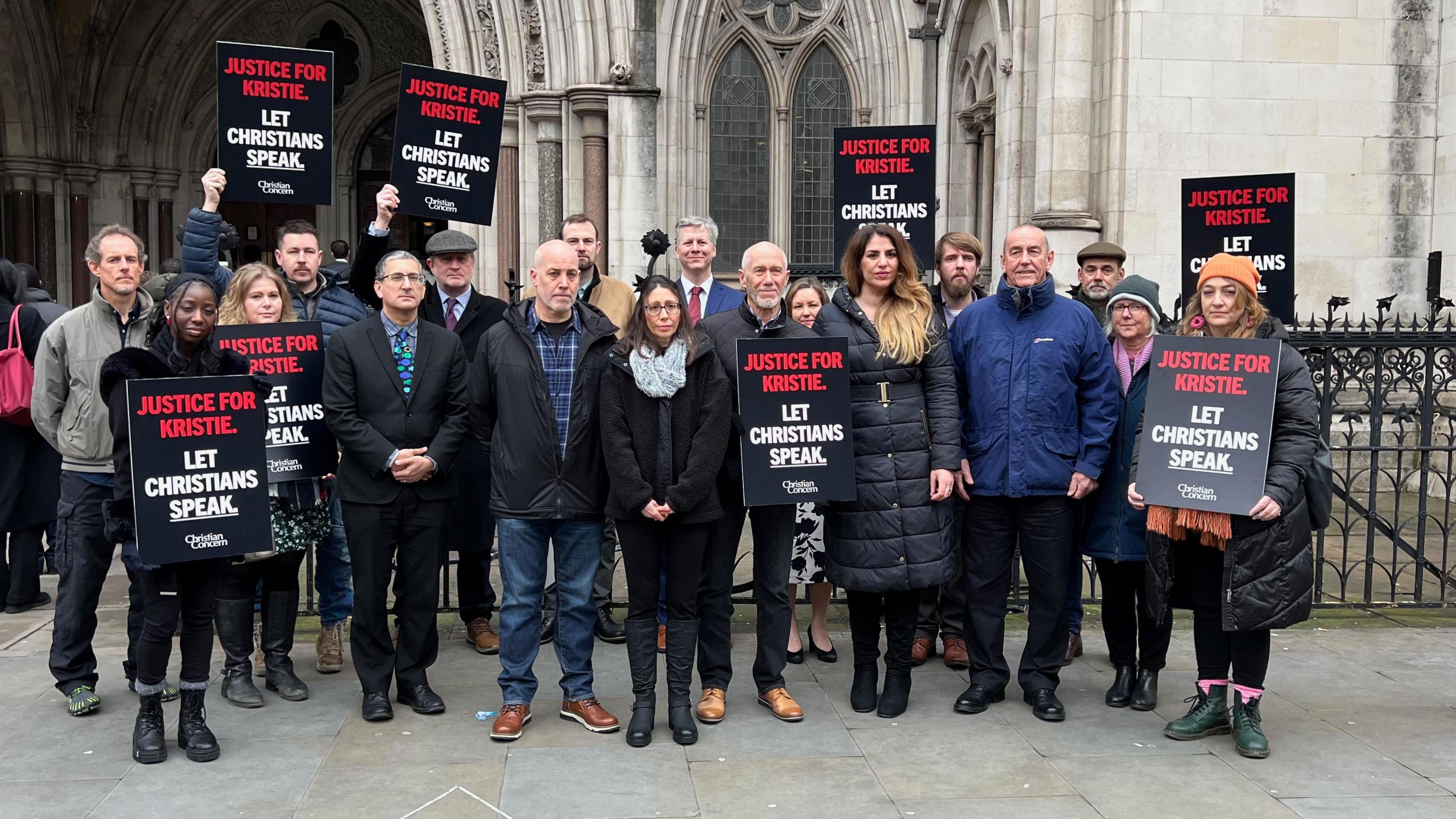 Kristie Higgs, surrounded by a group of supporters outside the High Court. Most are wearing winter coats and some are holding black placards which say 'Justice for Christie' on them in red letters. They also say 'Let Christians Speak' on them in white letters. 