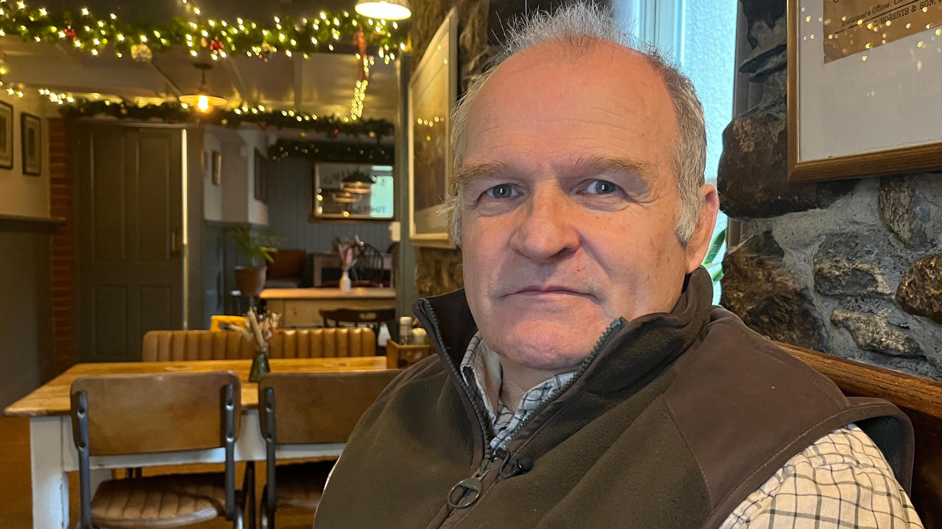 Llun o ddyn yn eistedd mewn bwyty yn edrych ar y camera. Mae ganddo wallt llwyd ac mae'n gwisgo crys siec gyda gilet brown. Tu ôl iddo mae bwrdd pren gyda phedair cadair o dan y bwrdd. Mae yna ddrws gwyrdd a goleuadau Nadolig ar y nenfwd.