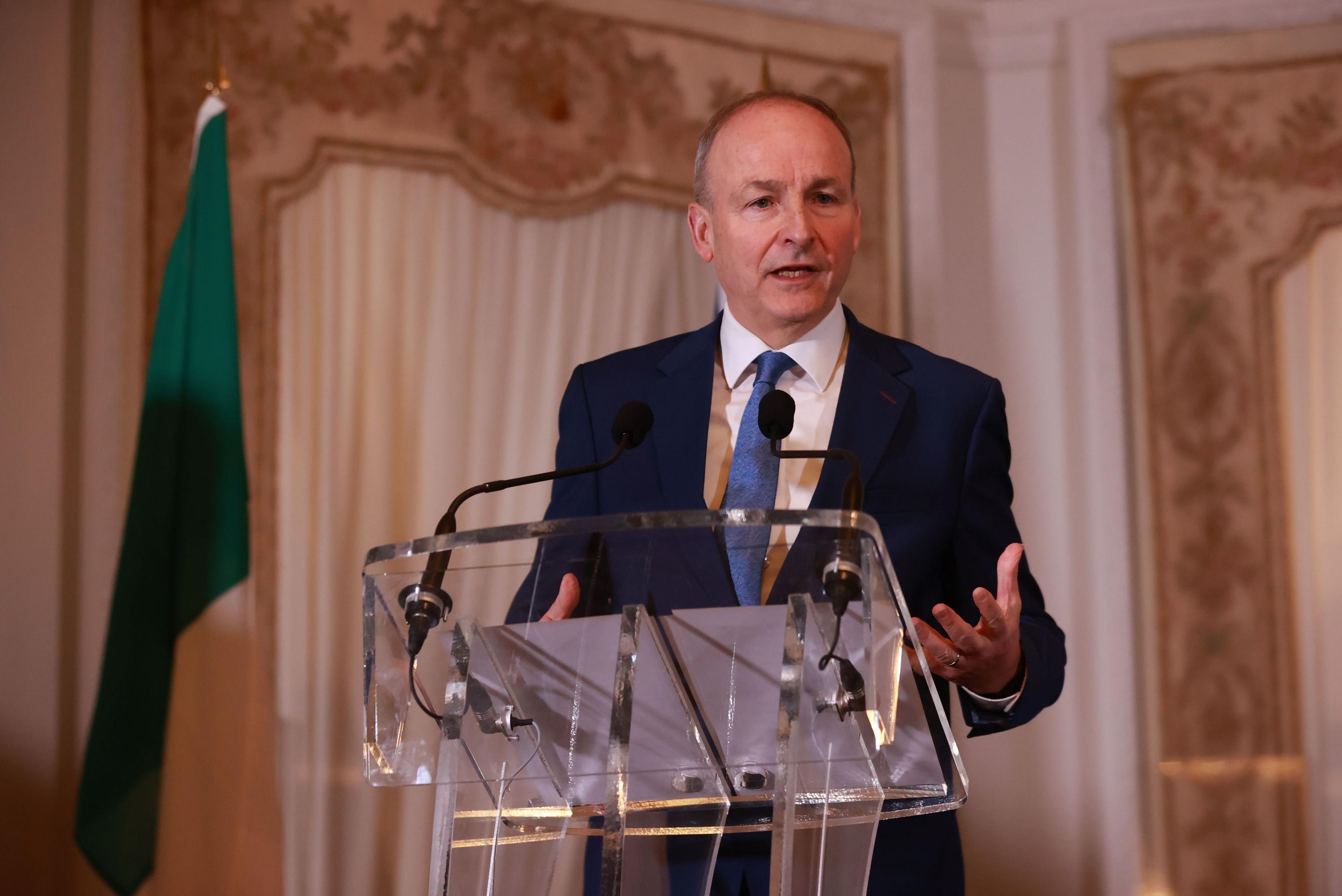 Fianna Fáil leader Micheal Martin is wearing a dark suit, white shirt and blue tie. He is standing behind a glass podium gesturing with his hands. An Irish flag can be seen furled in the background.